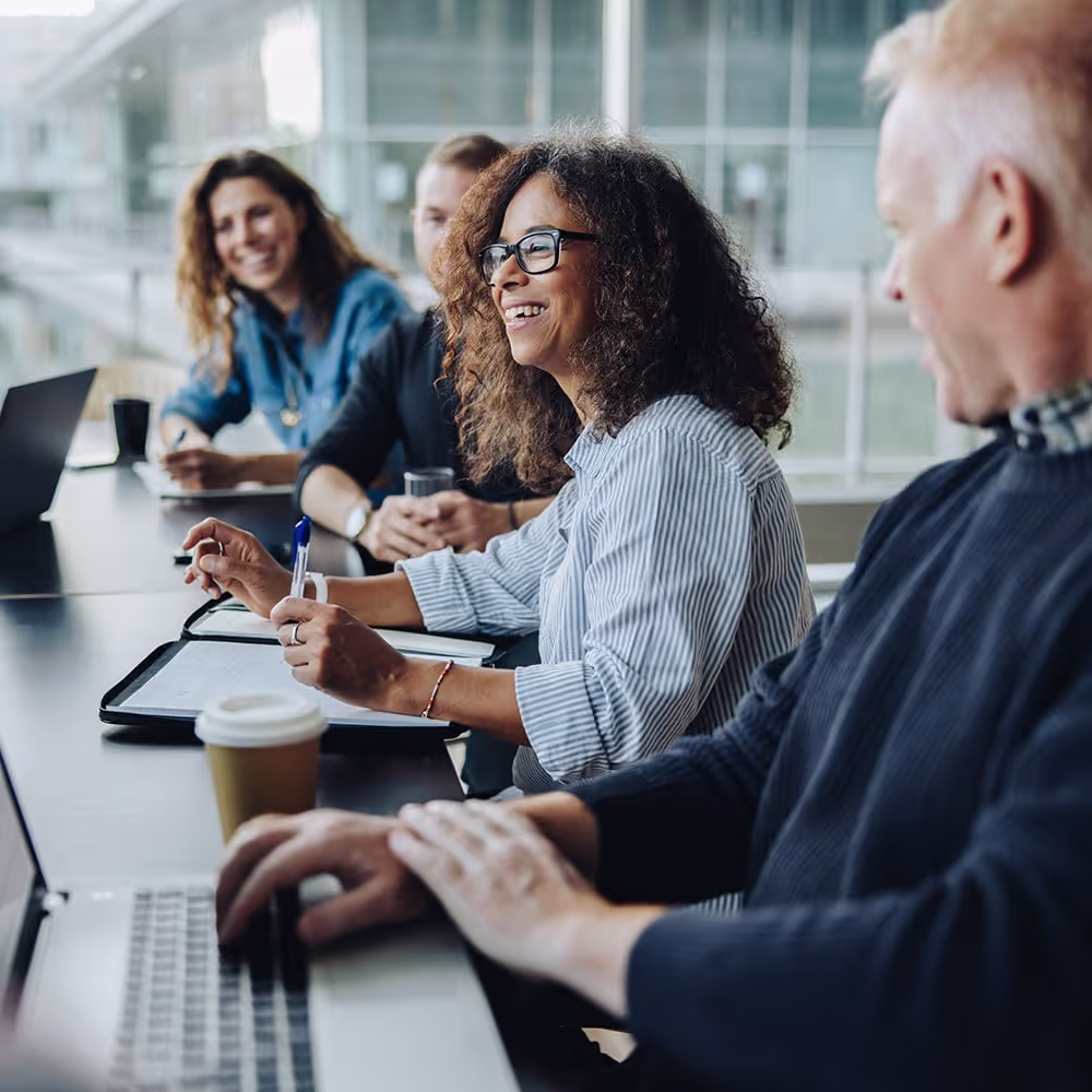 A group of professionals is engaged in a meeting, with a smiling woman taking notes while others participate in discussion.