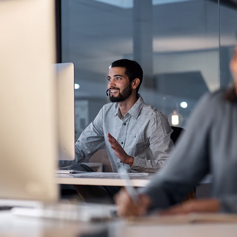 A man, seated at a desk in a modern office, is engaged in a conversation via a headset at night.