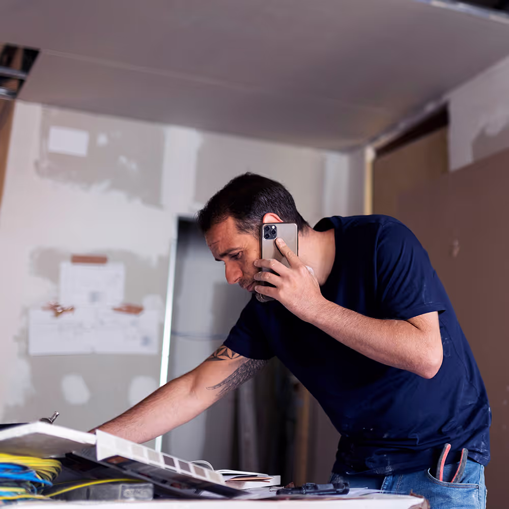 A man, likely a construction worker, bent over a work table in a room undergoing renovation talking on a smartphone.