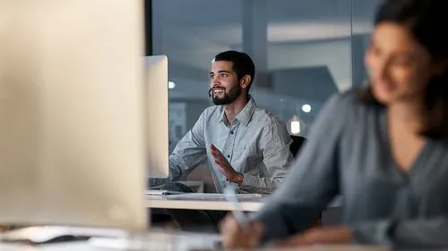 A man, seated at a desk in a modern office, is engaged in a conversation via a headset at night.