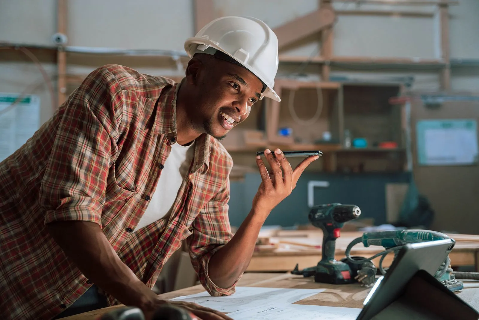 A man, likely a carpenter or construction worker, working in a workshop, he is speaking into a smartphone.