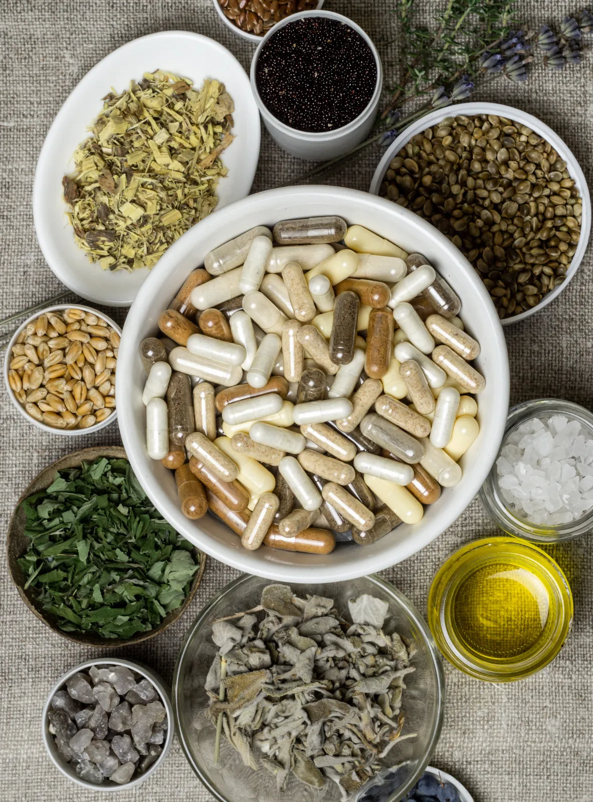 Bowl of supplement capsules surrounded by dried botanicals, seeds and oil in small dishes