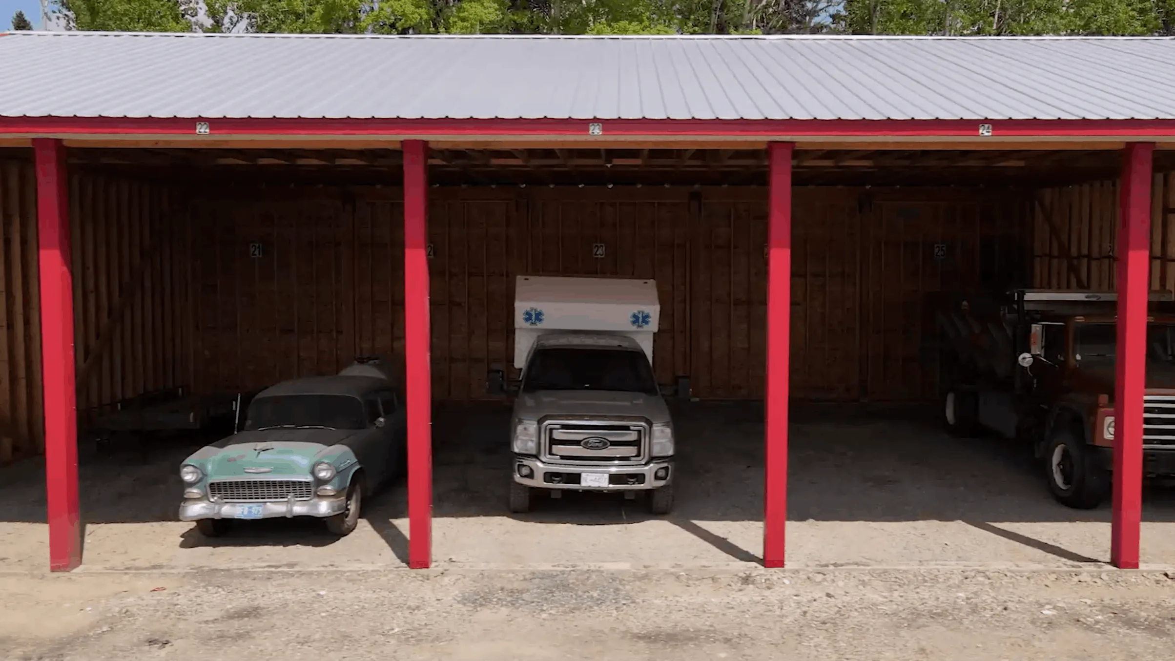 Three vehicles parked under a metal-roof shelter with red support beams, including a vintage turquoise station wagon, a silver ambulance truck, and a brown flatbed truck.