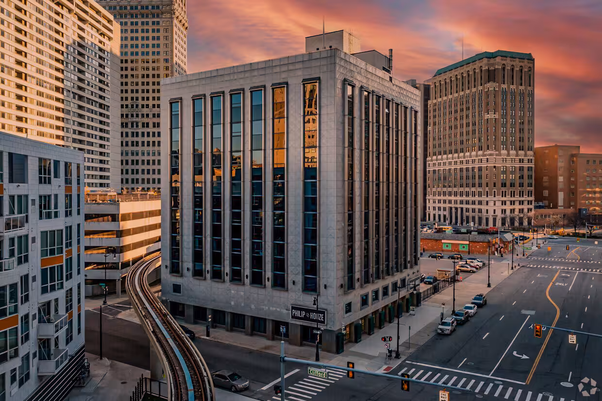 Urban street intersection with a mid-century apartment building in Downtown Detroit reflecting sunset colors, adjacent railroad tracks, and surrounding city buildings under a vibrant evening sky.