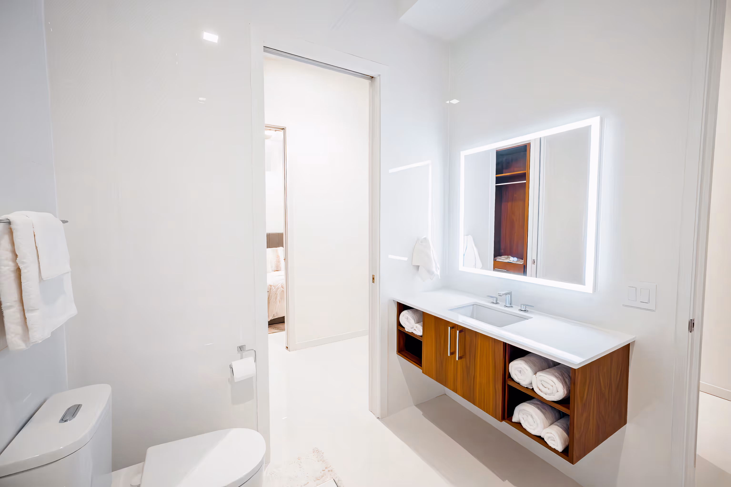 Bright white bathroom with a floating wooden vanity, illuminated rectangular mirror, toilet, and white towels.