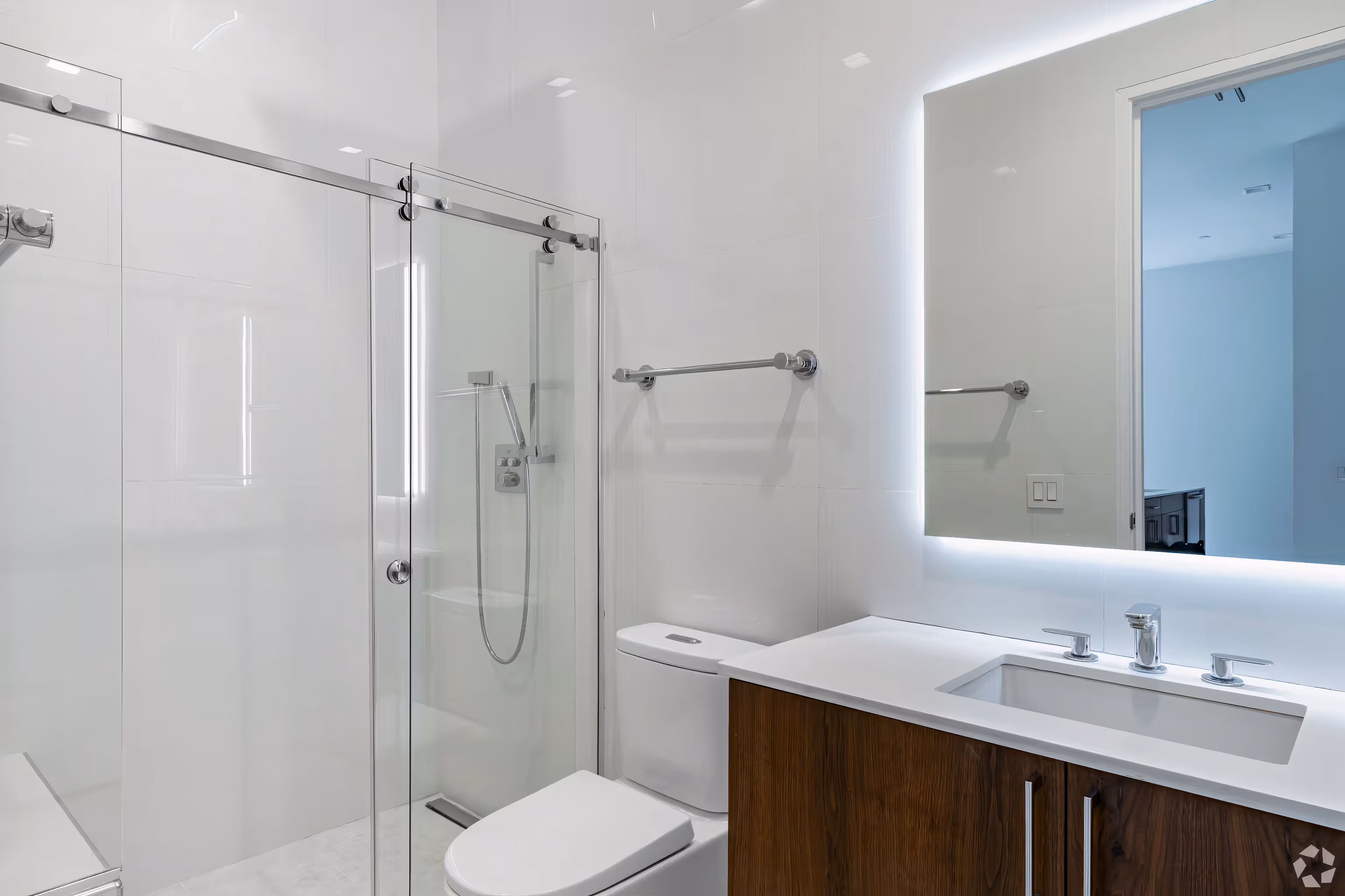 Modern bathroom with glass-enclosed shower, white toilet, wood vanity, and backlit mirror.