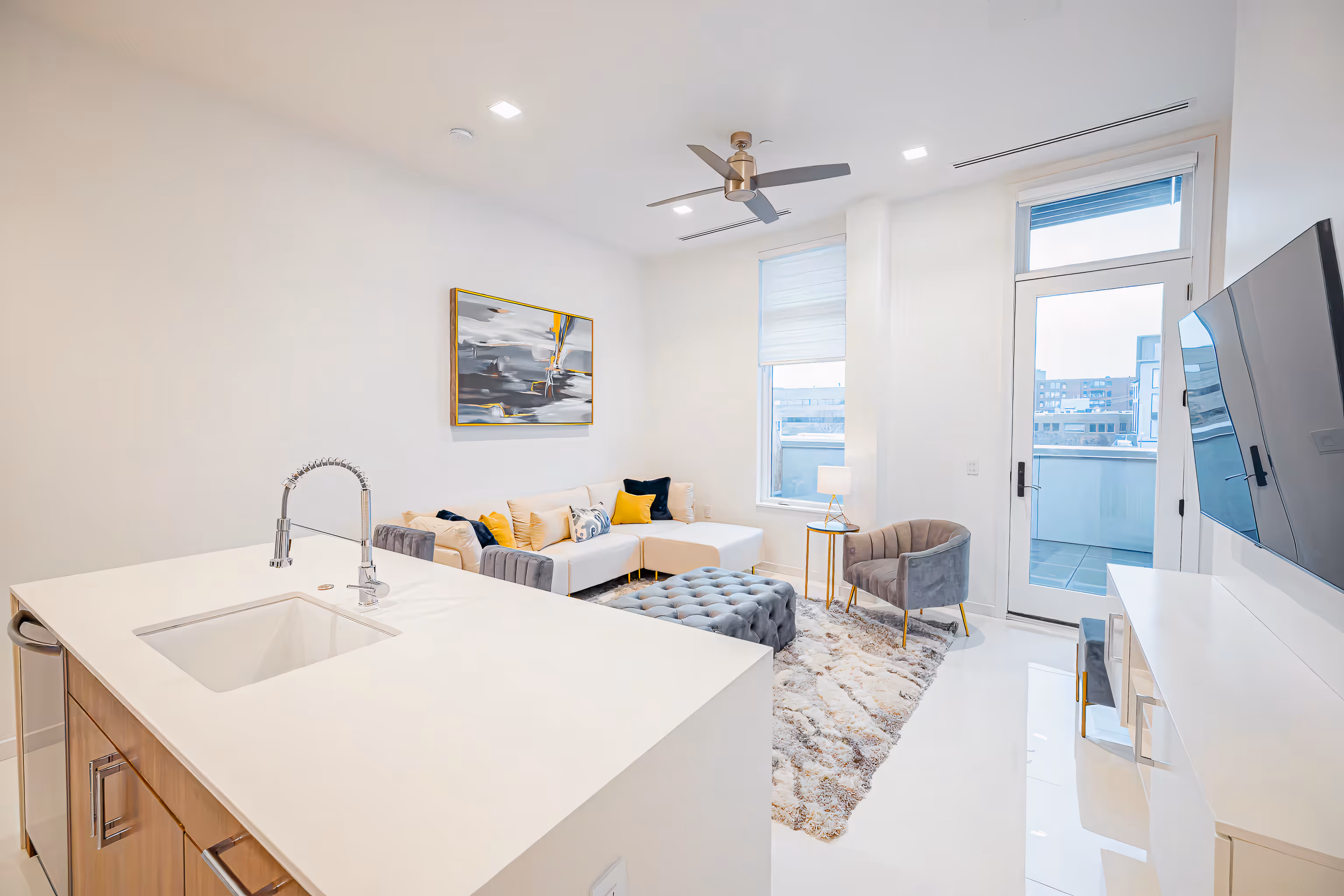 Bright modern living room with white sectional sofa, gray armchair, tufted ottoman, abstract painting, and a kitchen island with sink.