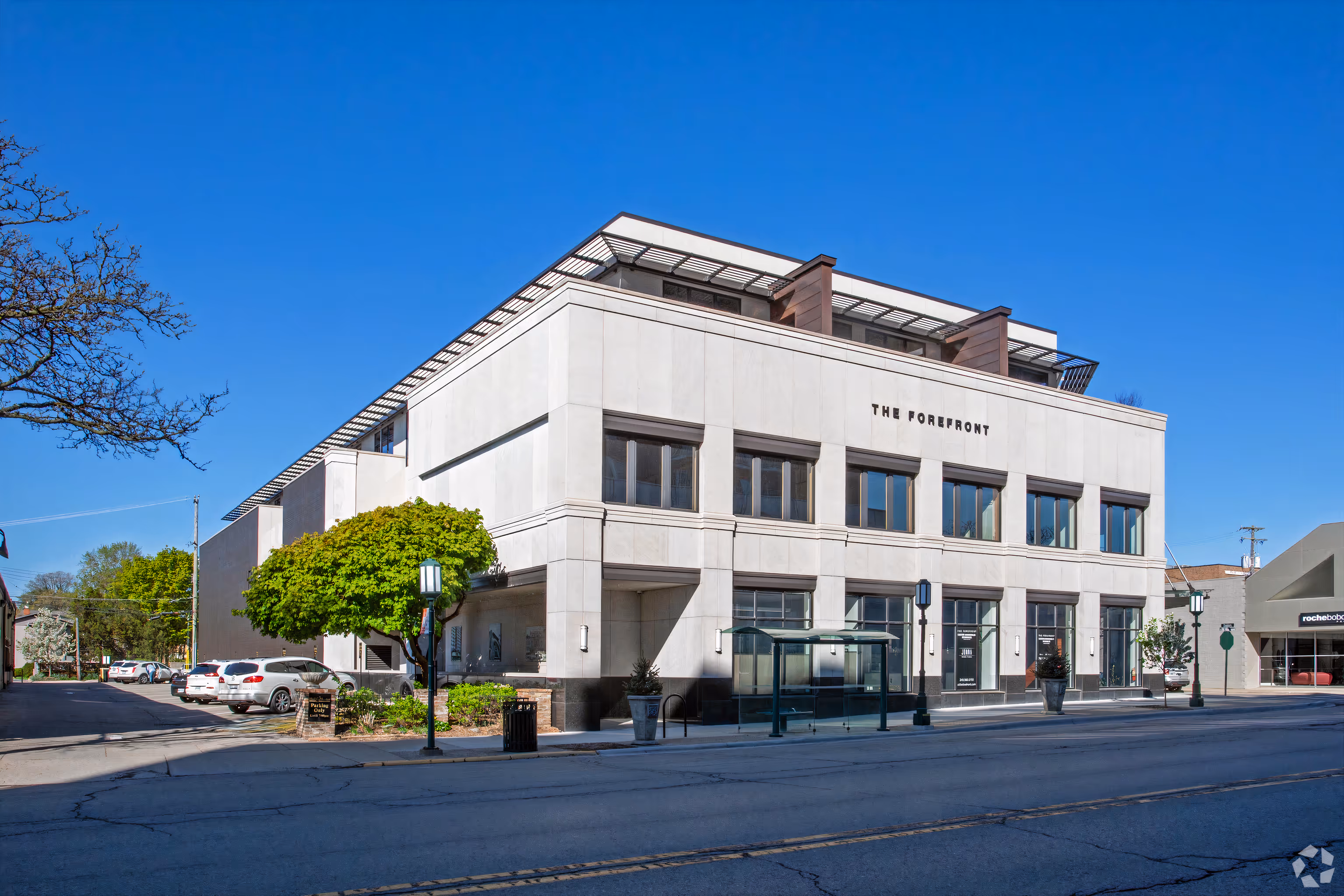 Luxury two-story apartment building named The Forefront with large windows, a bus stop shelter, and a tree-lined street under a clear blue sky.