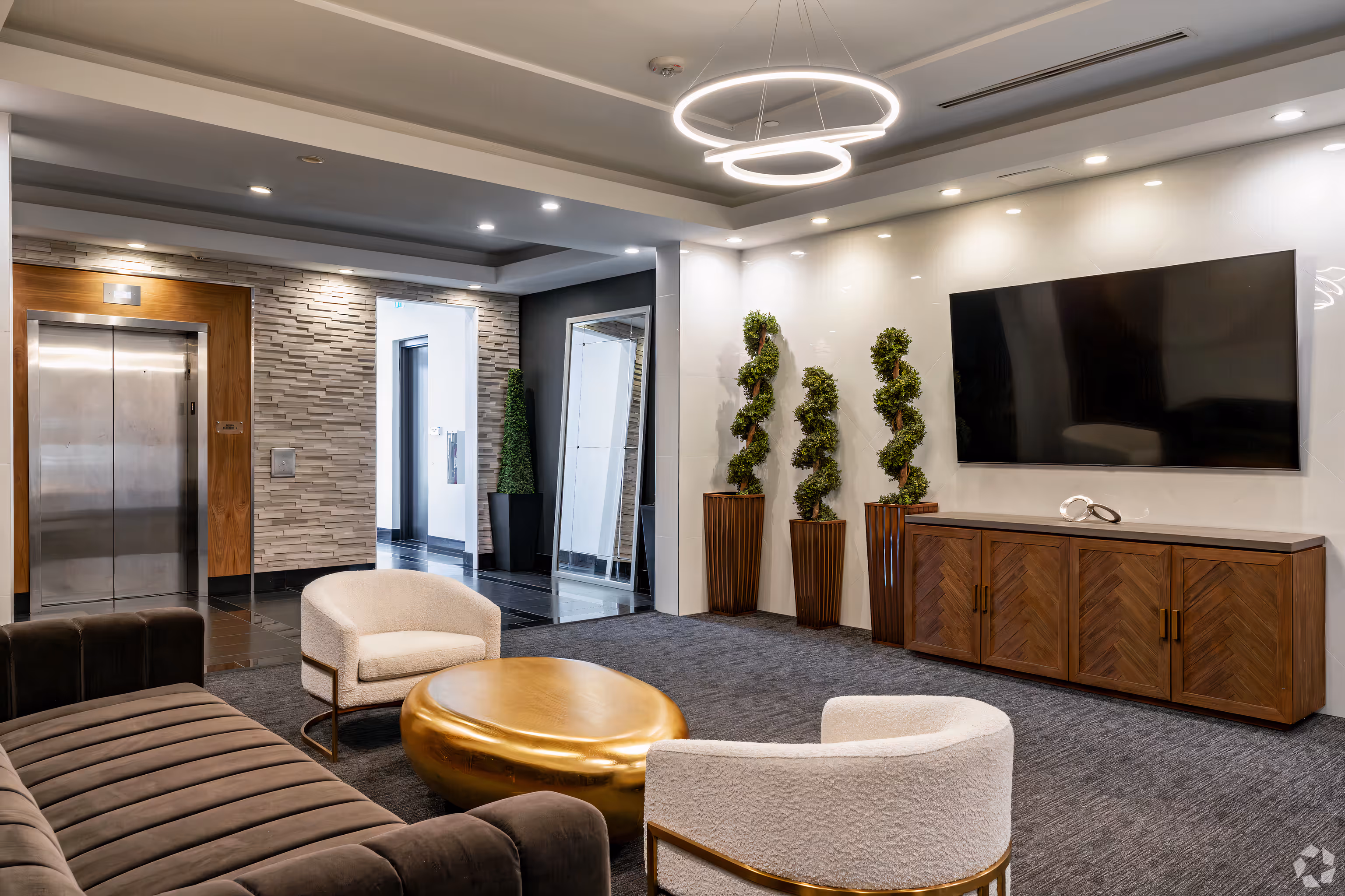 Modern lounge area with plush chairs, gold round coffee table, wall-mounted TV above wooden cabinet, and elevator in the background.