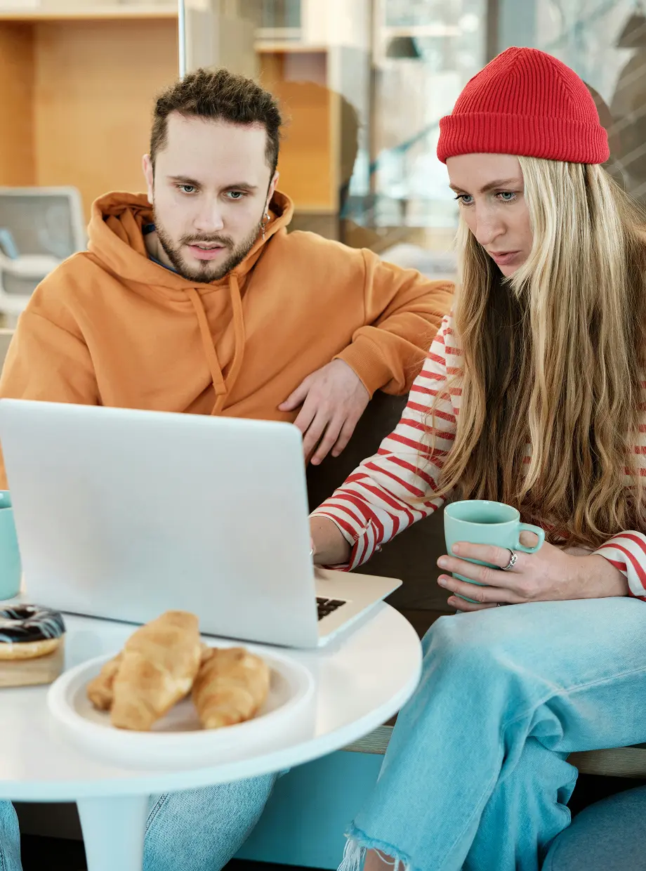 Two people sitting at a table with a laptop, one wearing an orange hoodie and the other a red beanie holding a mug, with pastries on the table.