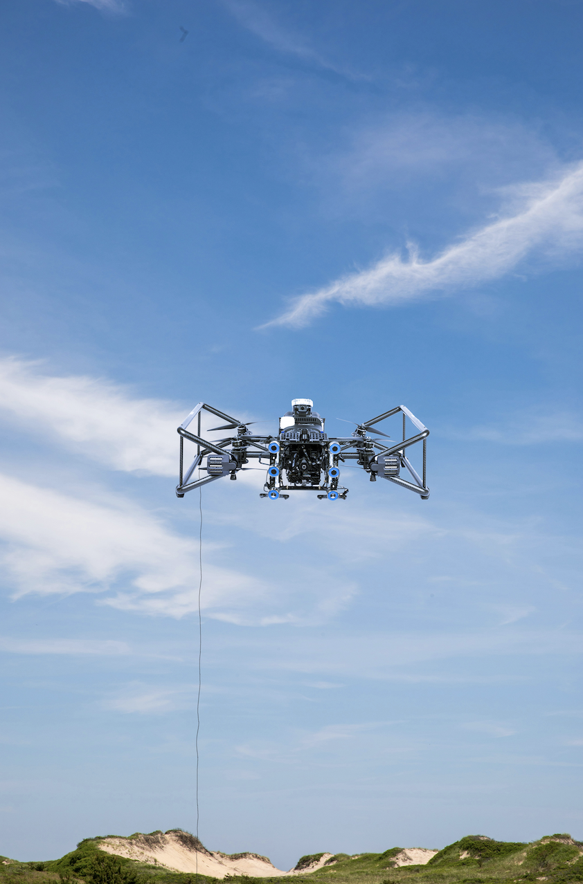 Black drone with cameras and sensors hovering over grassy dunes under a blue sky with wispy clouds.
