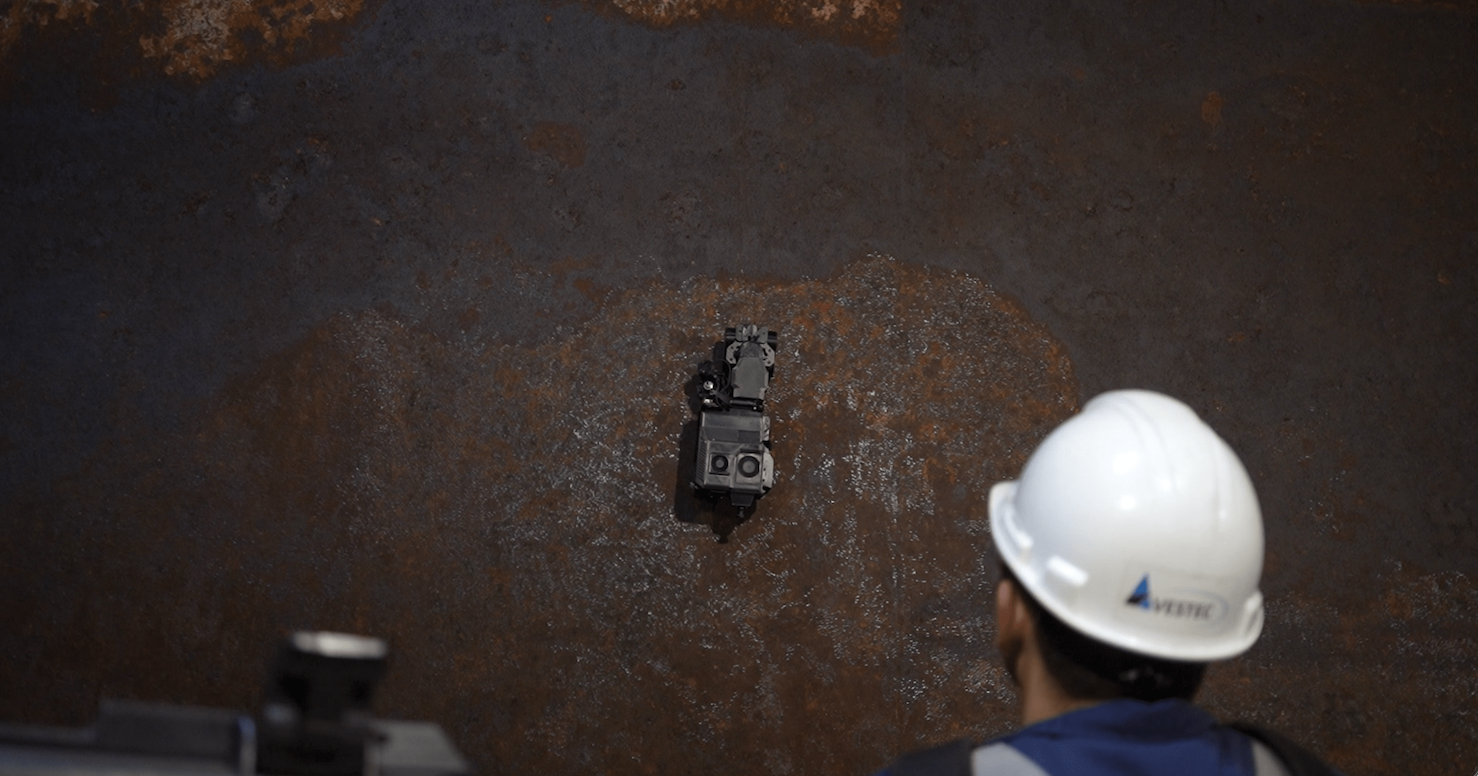 Person wearing a white hard hat inspecting a small robotic device on a rusty metal surface.