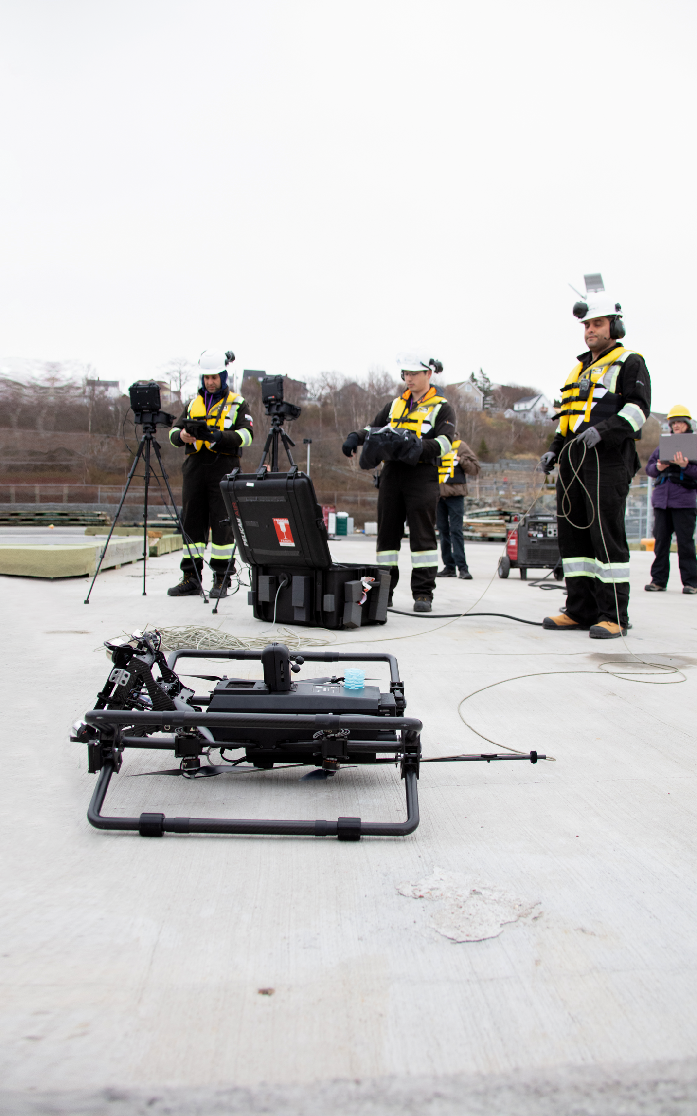 Technicians in safety gear operate a drone and equipment on a concrete surface outdoors.