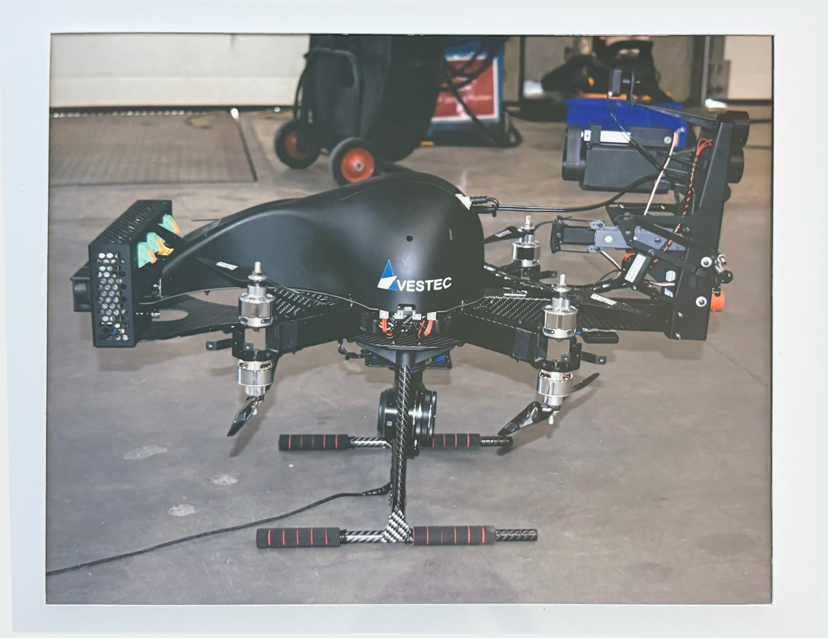 Black Vestec drone with four rotors, carbon fiber parts, and a camera system mounted underneath, on a concrete floor indoors.