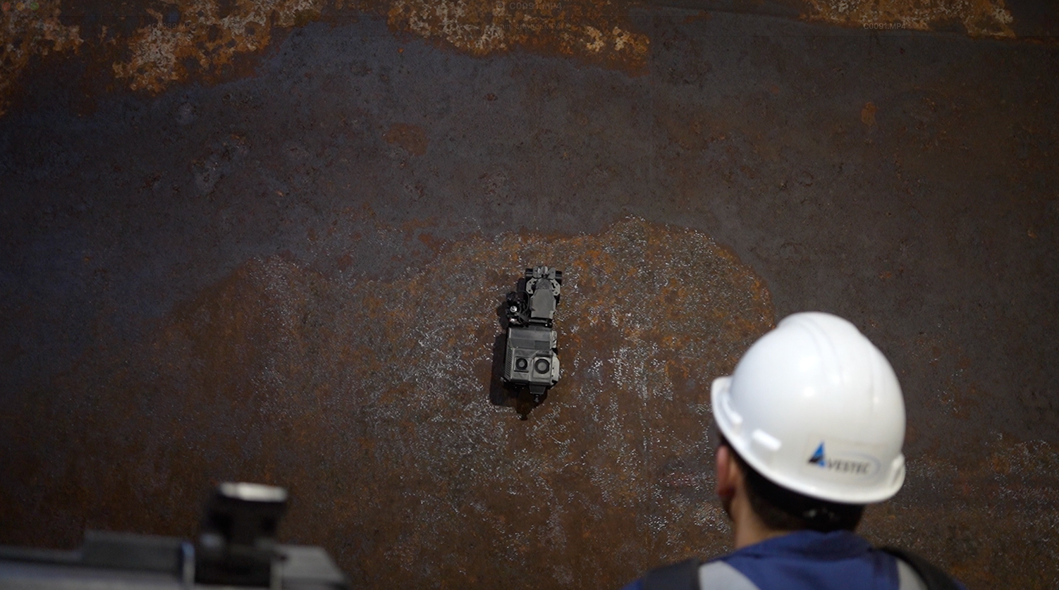 Person wearing a white hard hat inspecting a small device mounted on a large rusted metal surface.