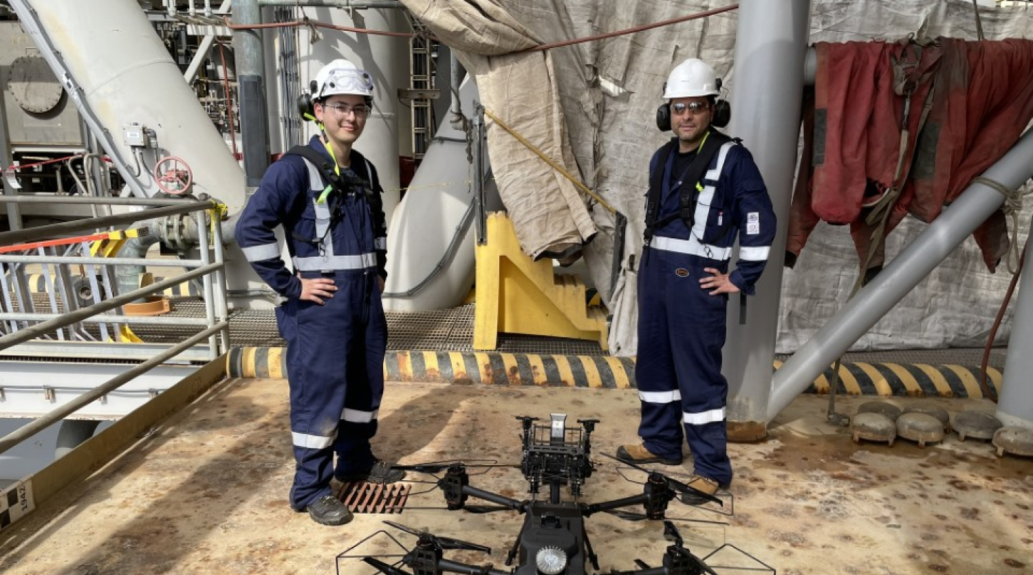 Avestec staff wearing navy blue protective coveralls and white helmets standing on a metal platform with a large black drone on the ground between them.
