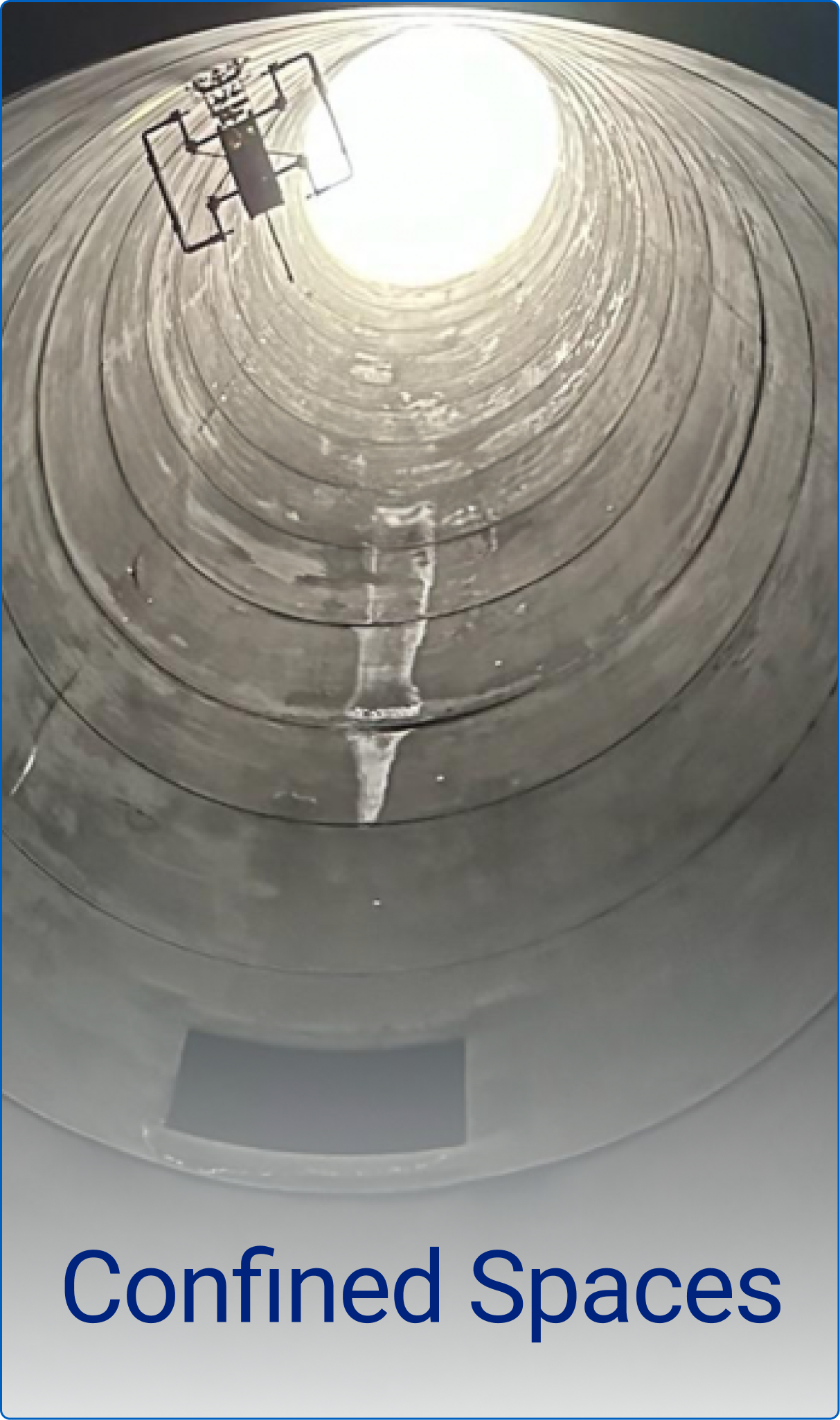 View looking upward inside a tall cylindrical concrete confined space with sunlight at the opening and a safety cage ladder on the side.