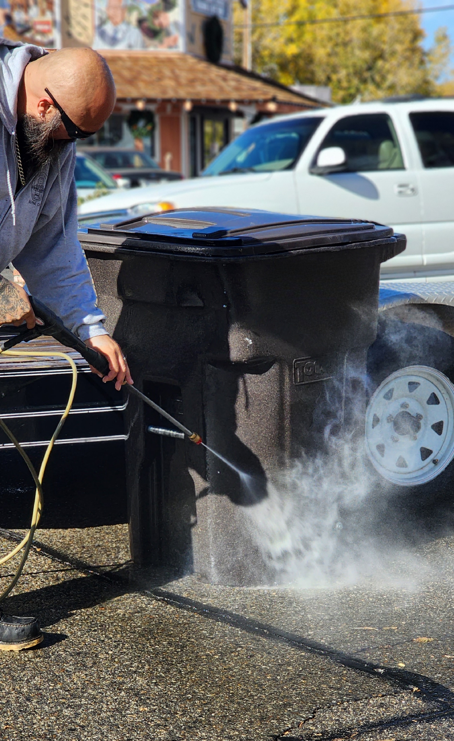 Man with sunglasses pressure washing a black trash bin on a sunny street.