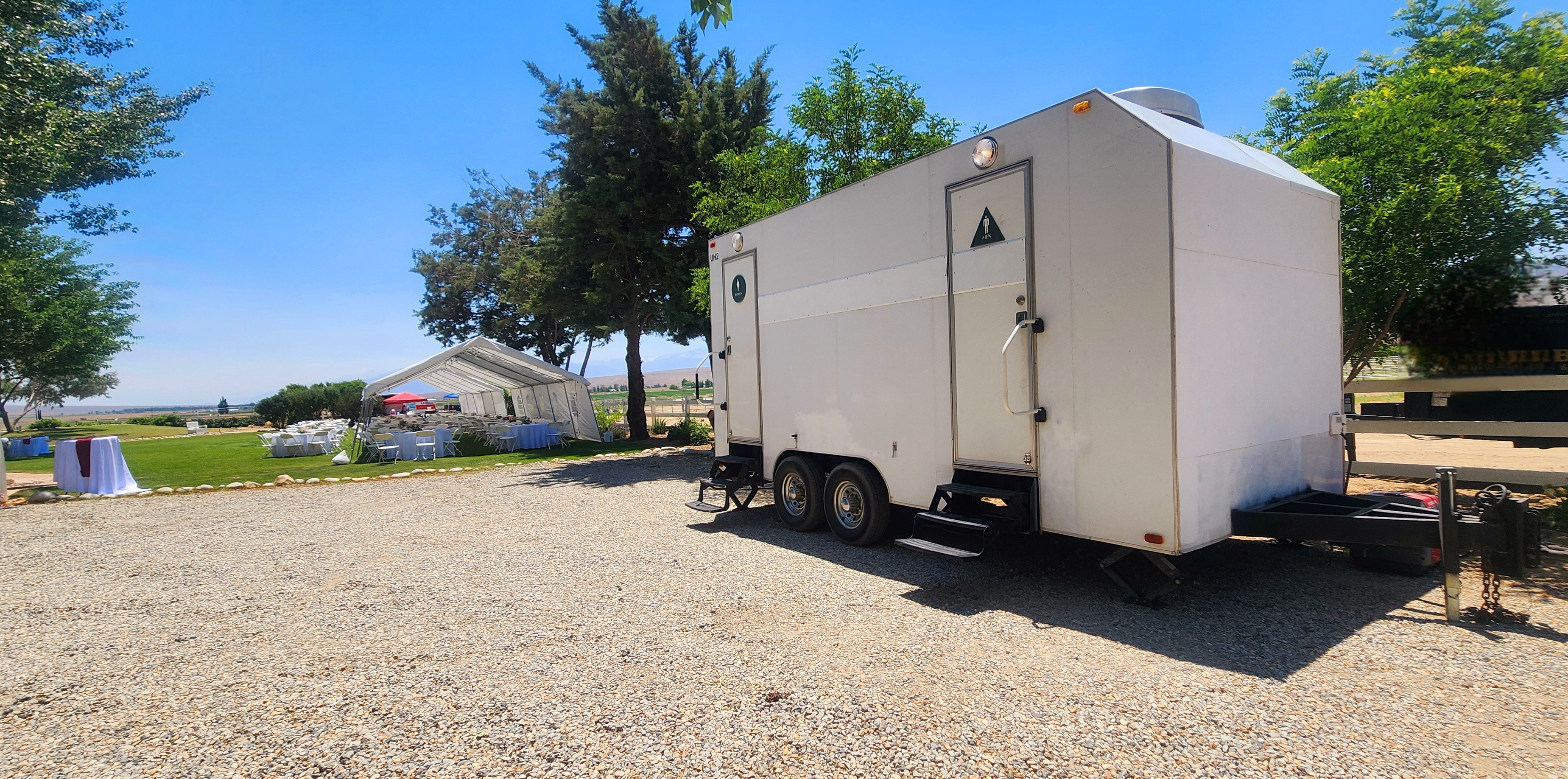 White portable restroom trailer with men’s and women’s doors parked on gravel near outdoor event tents with tables and chairs on a sunny day.