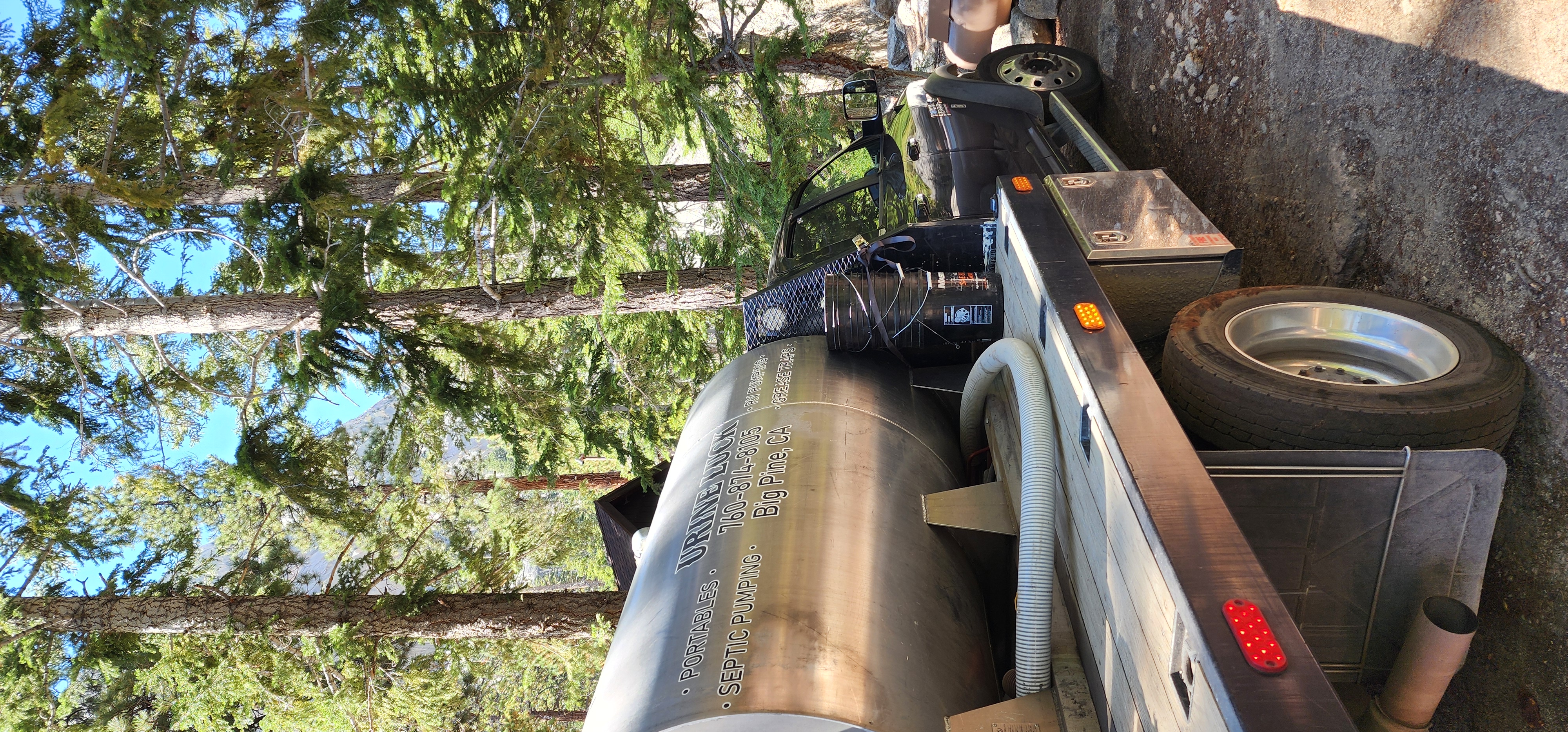 Flatbed truck with a large metal septic tank and hoses parked in a forested area with tall pine trees.