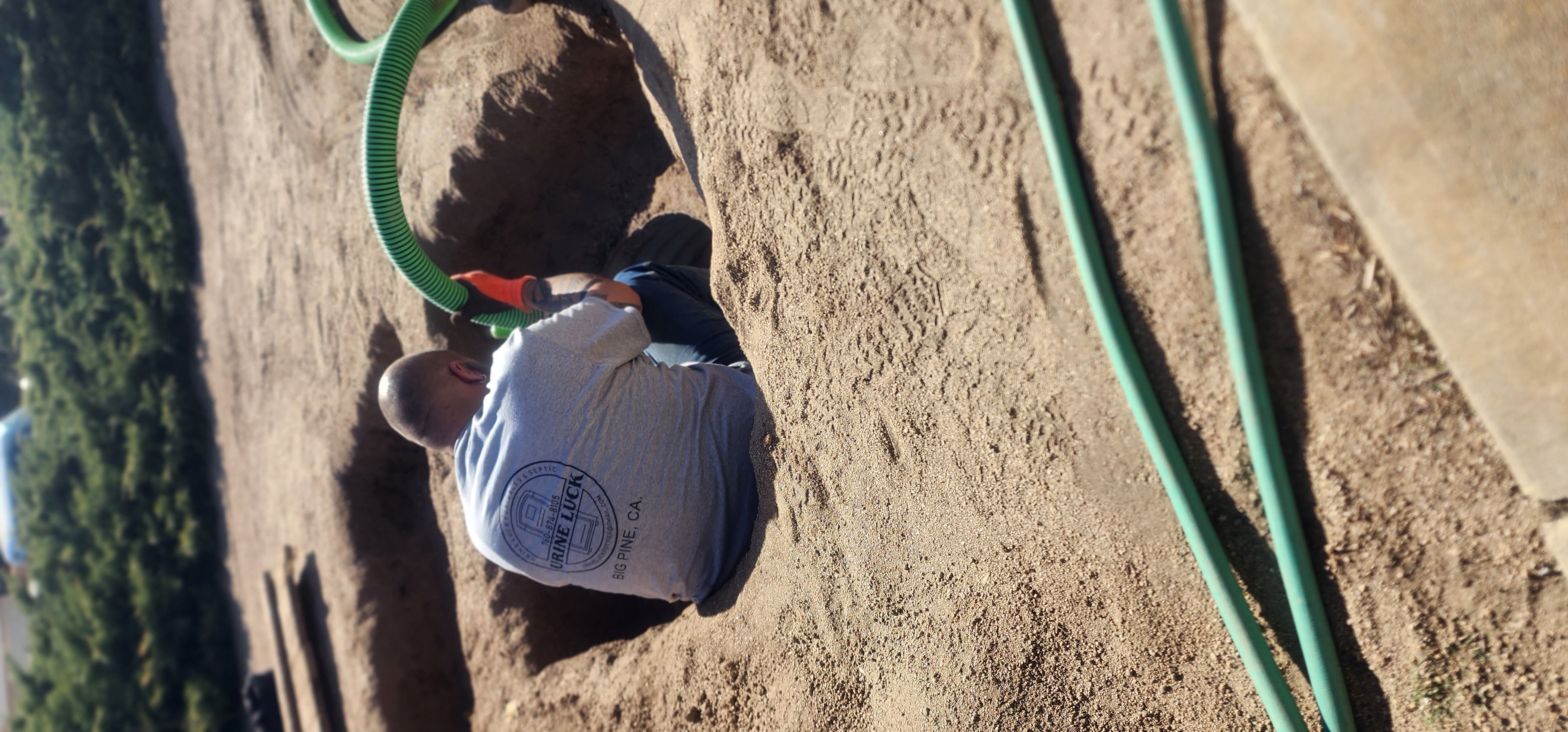 Man wearing a gray shirt and orange glove sitting in a dirt hole holding a green flexible hose.