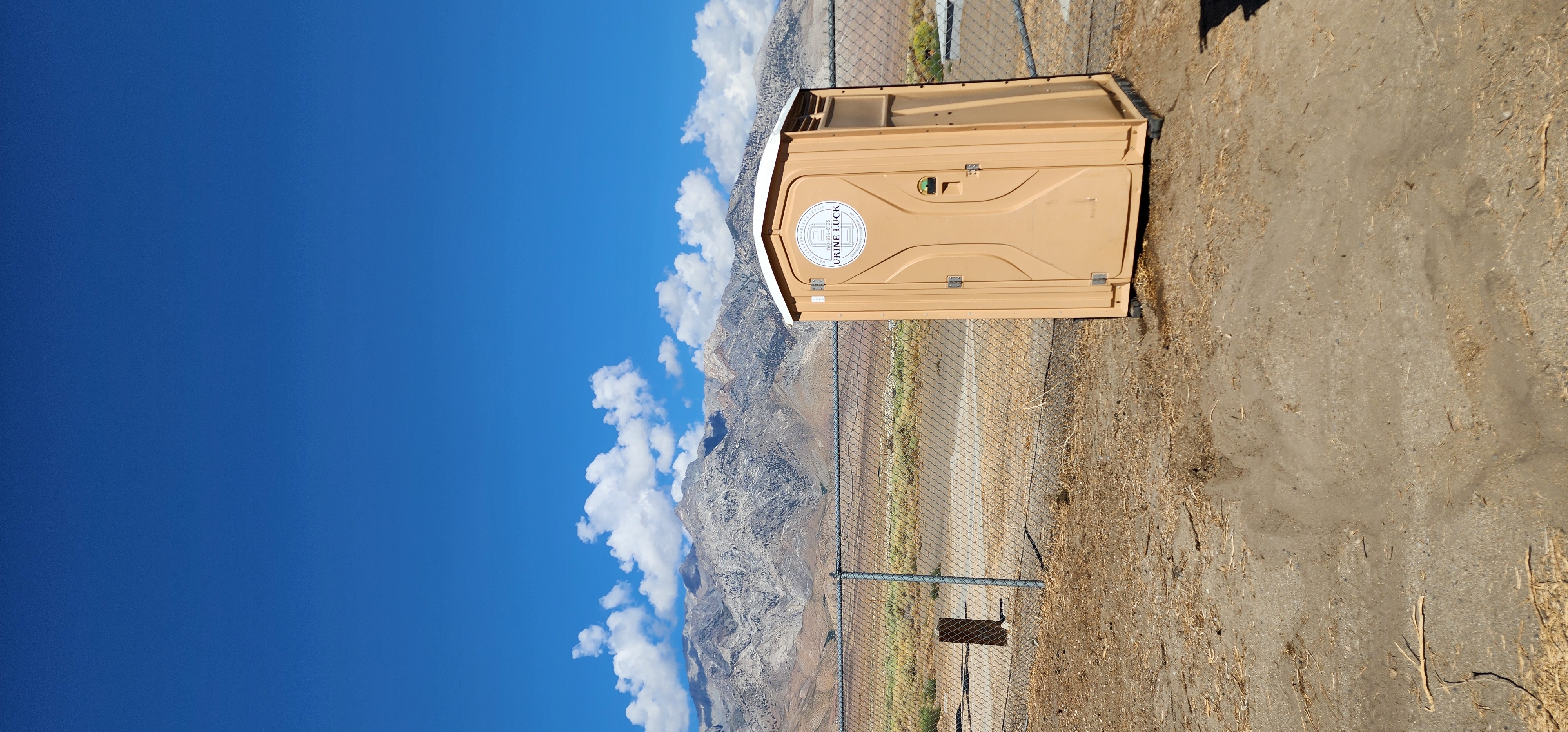 Beige portable toilet standing on dry ground near a chain-link fence with mountains and a blue sky with clouds in the background.