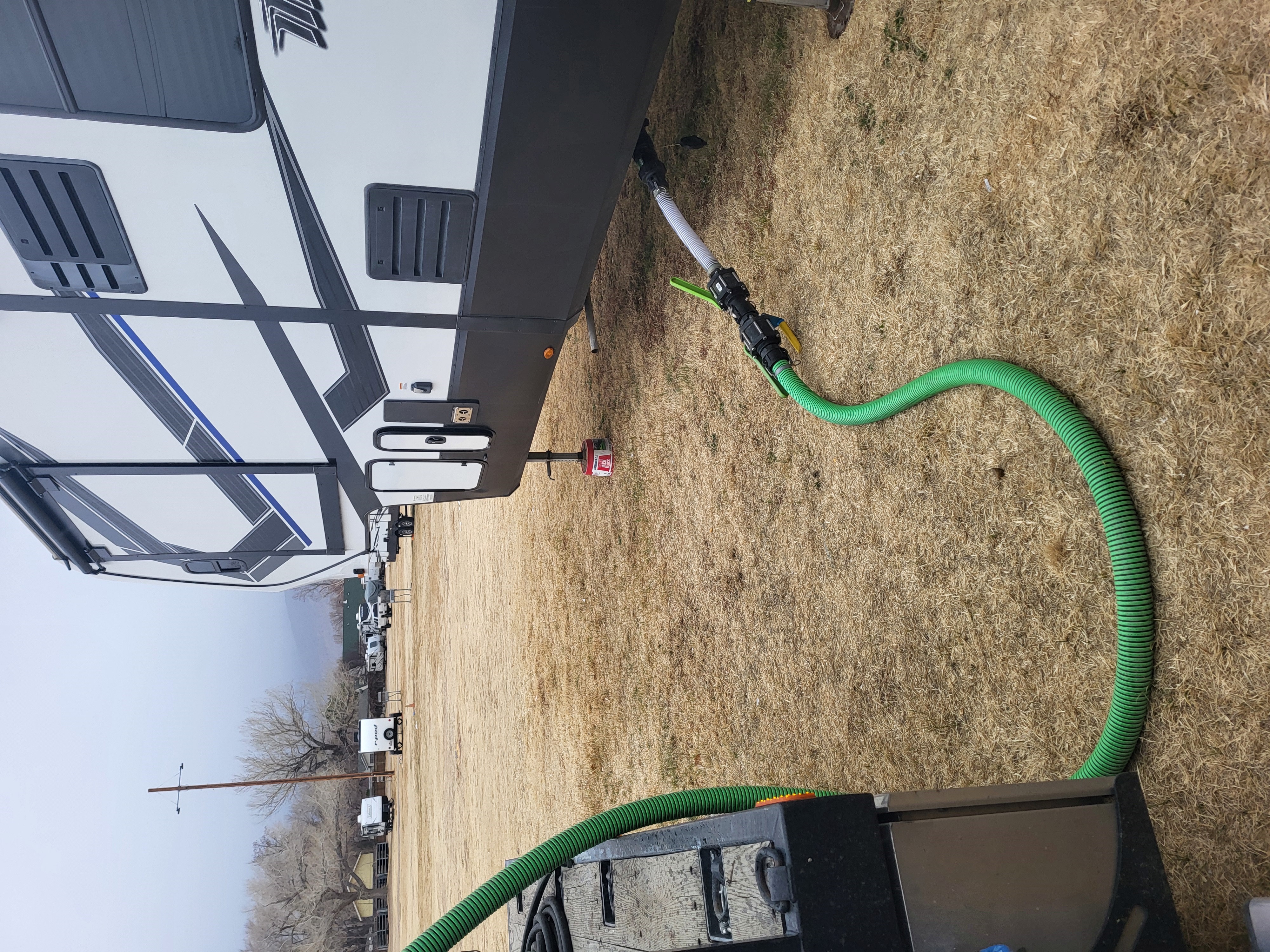 Green hose connected to the side of a parked white and black RV on dry grass with other RVs and leafless trees in the background.