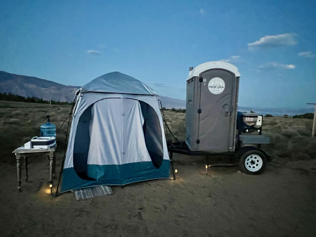 Portable restroom trailer parked next to a small tent and a table with a water jug in a rural outdoor setting at dusk.