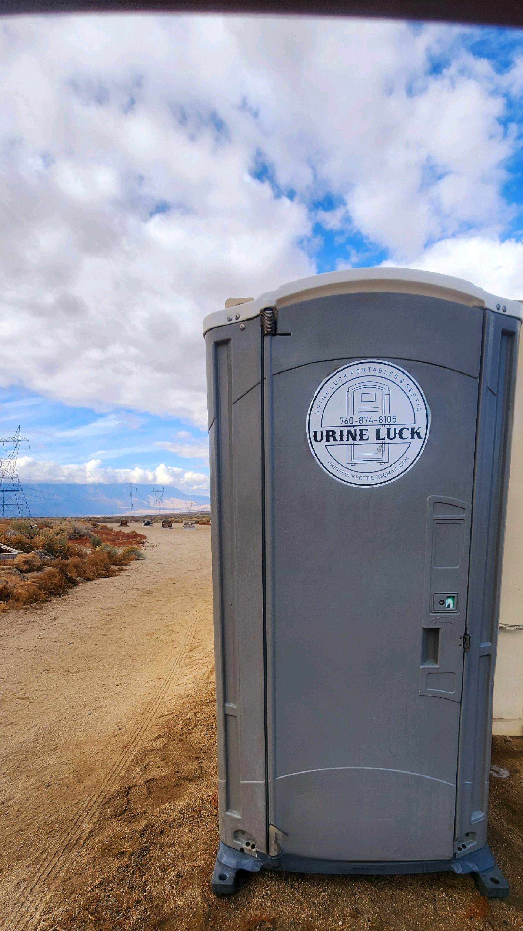 Gray portable toilet with a Urine Luck company logo standing on dirt road in a desert landscape under a cloudy blue sky.
