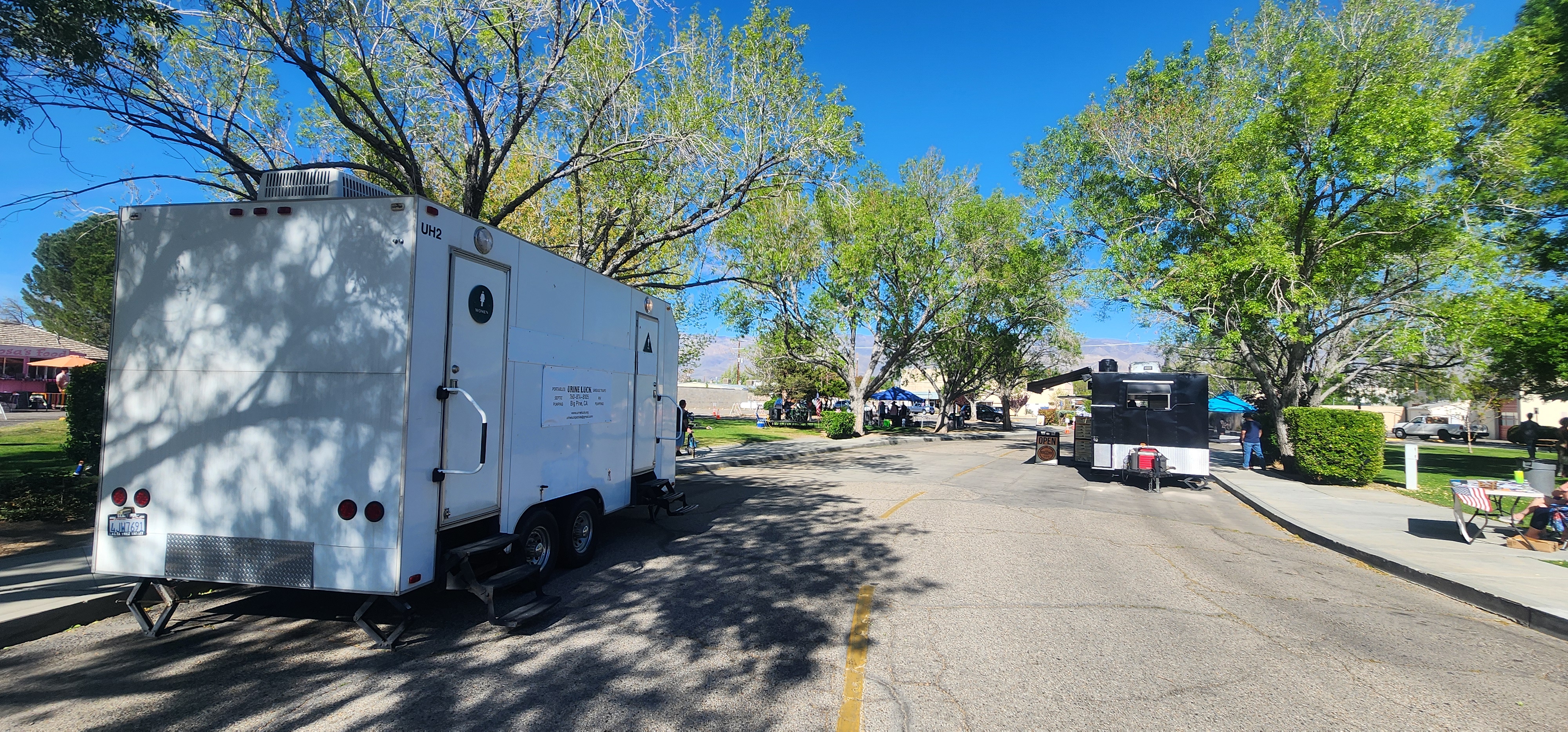 White portable restroom trailer and black food truck parked on a street near green trees and a park on a sunny day.