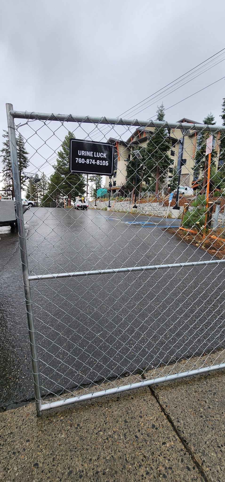 Chain-link fence gate with a black sign reading 'URINE LUCK 760-874-8105' blocking a wet paved road near a building and trees under an overcast sky.