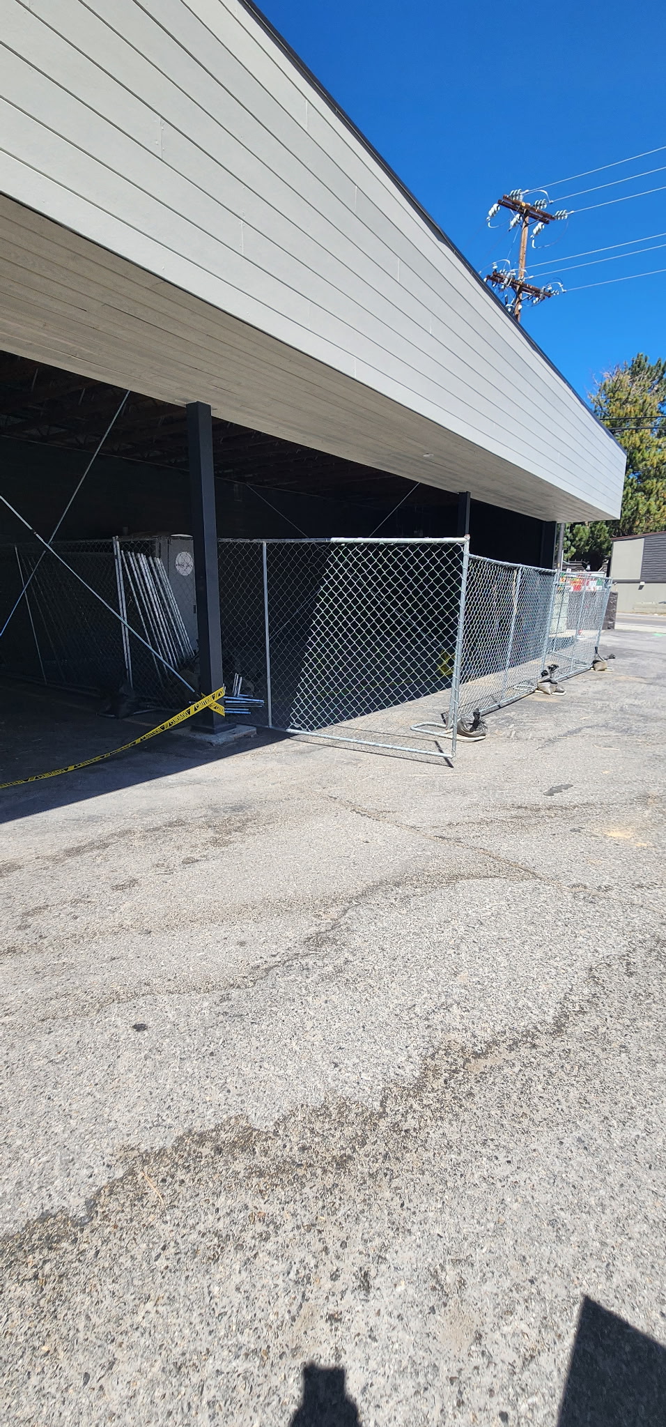 Metal chain-link fence with yellow caution tape enclosing a shaded area beneath a building overhang on a sunny day.