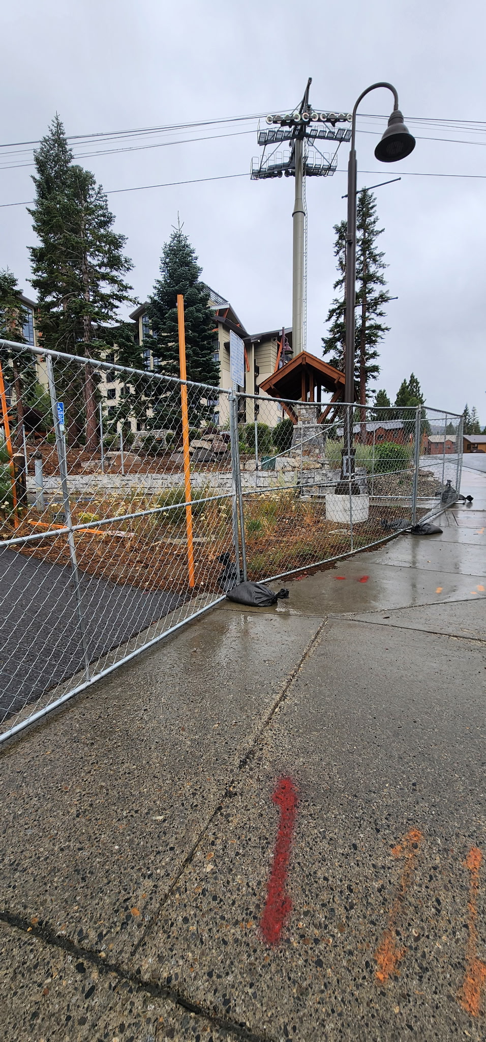 Chain-link construction fence on wet sidewalk with red arrow painted on concrete, pine trees, lamp post, and buildings in the background under cloudy sky.