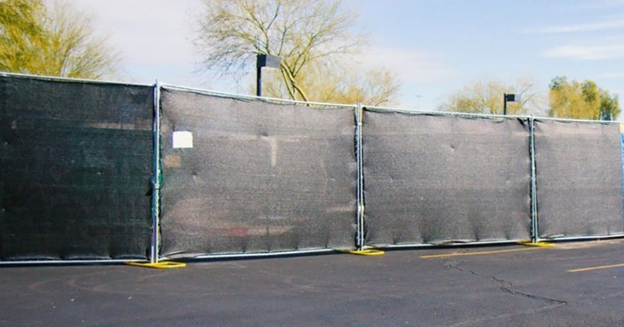 Black mesh privacy fence panels set up on an asphalt surface with trees and sky in the background.
