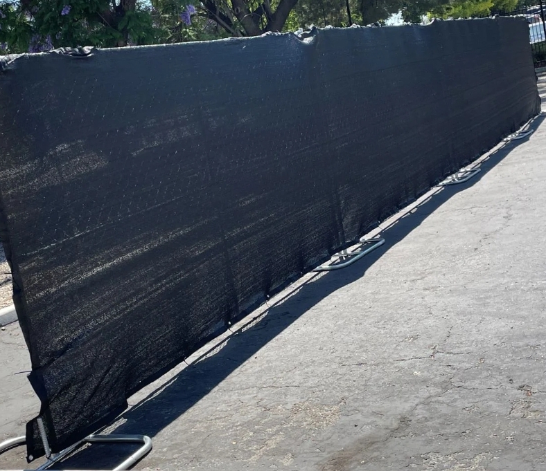 Long black mesh privacy screen secured to metal frame stands on a paved surface with trees and greenery in the background.