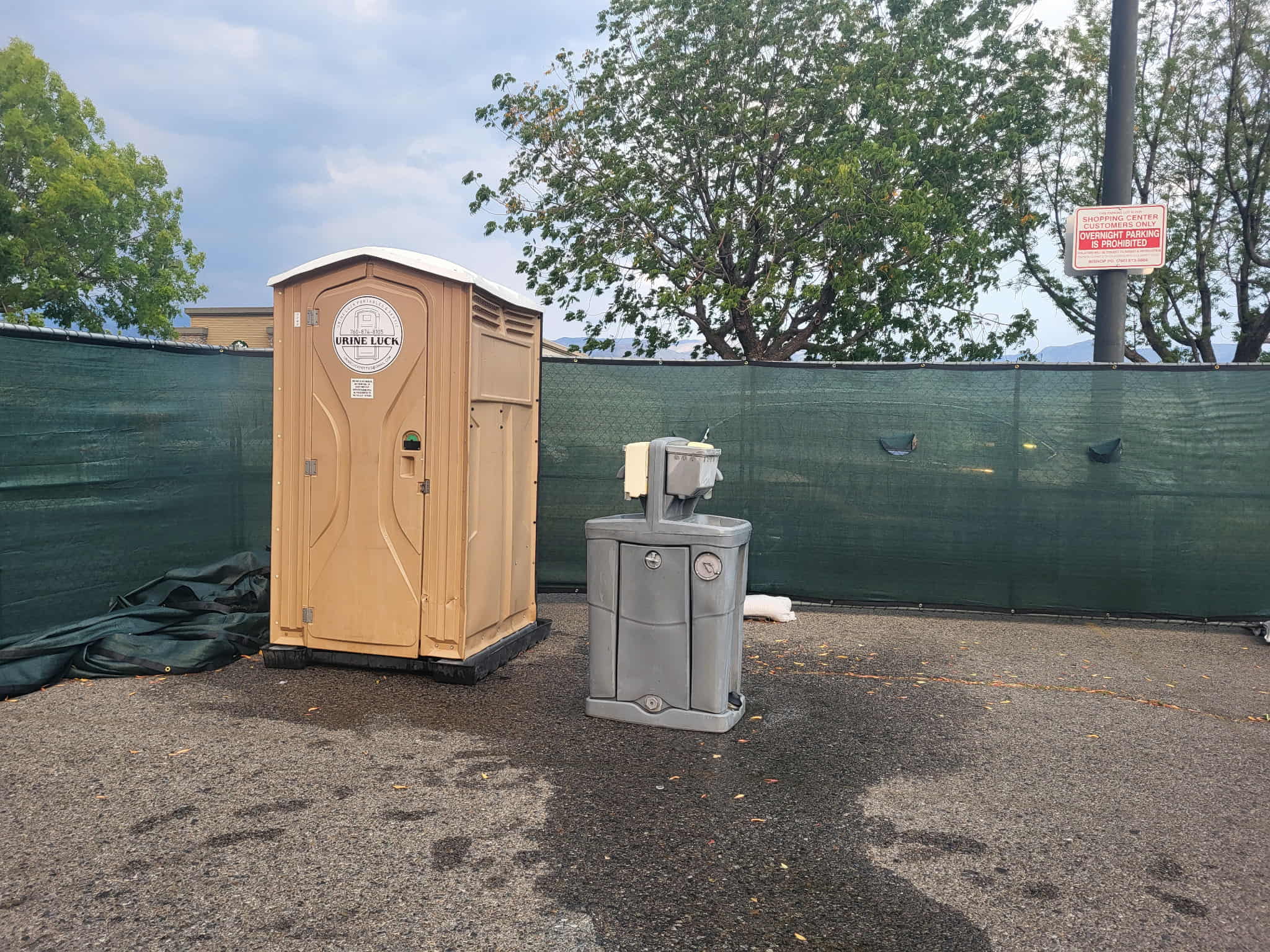 Portable beige toilet and gray water fountain on asphalt with green mesh fence and trees in the background.