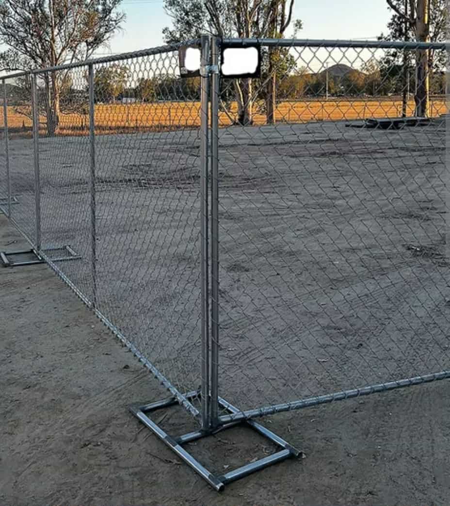 Temporary chain-link fence panels set up on a dirt ground with trees and a field in the background.