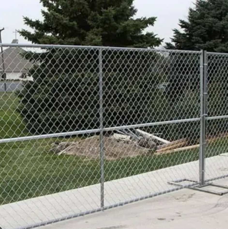 Temporary chain-link fence enclosing a grassy area with stacked construction materials and large evergreen trees in the background.
