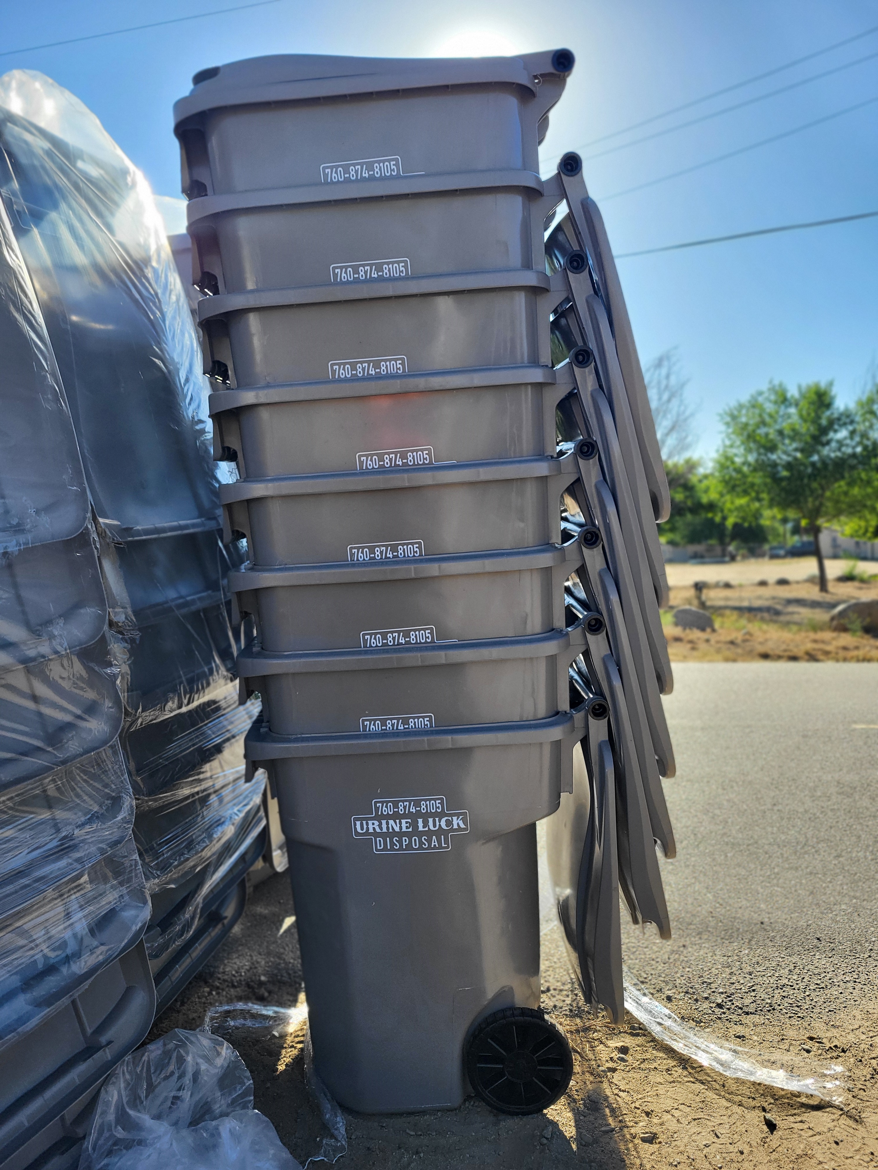 Stack of gray wheeled trash bins labeled 'Urine Luck Disposal' with phone number, outdoors on a sunny day.