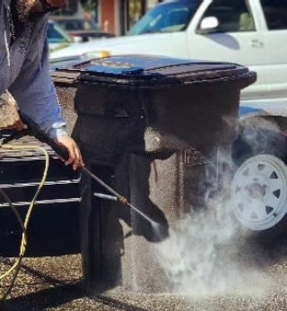 Person using a pressure washer to clean a black trash bin outdoors near a white vehicle.