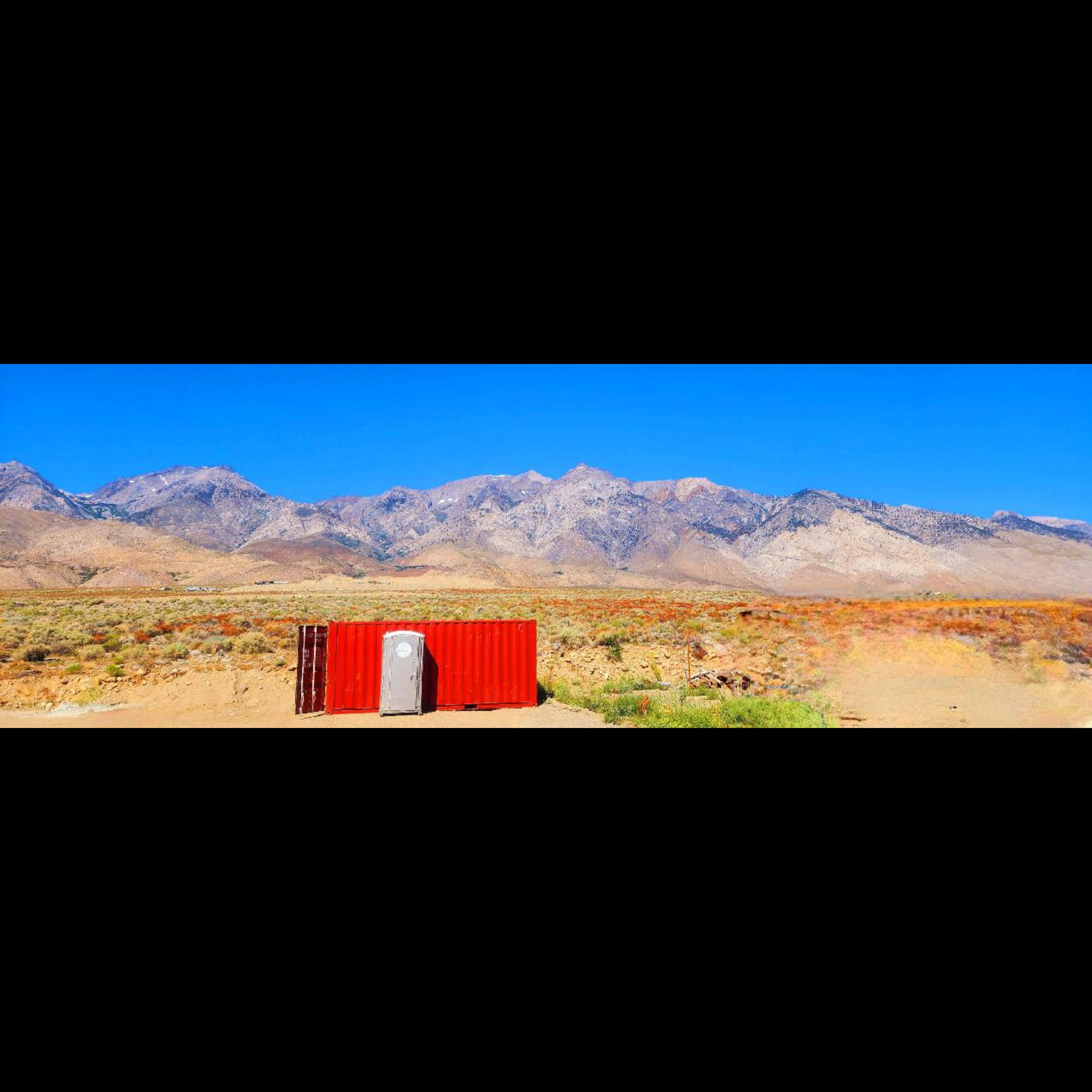 Red shipping container with a portable toilet in front, set in a desert landscape with mountains under a clear blue sky.