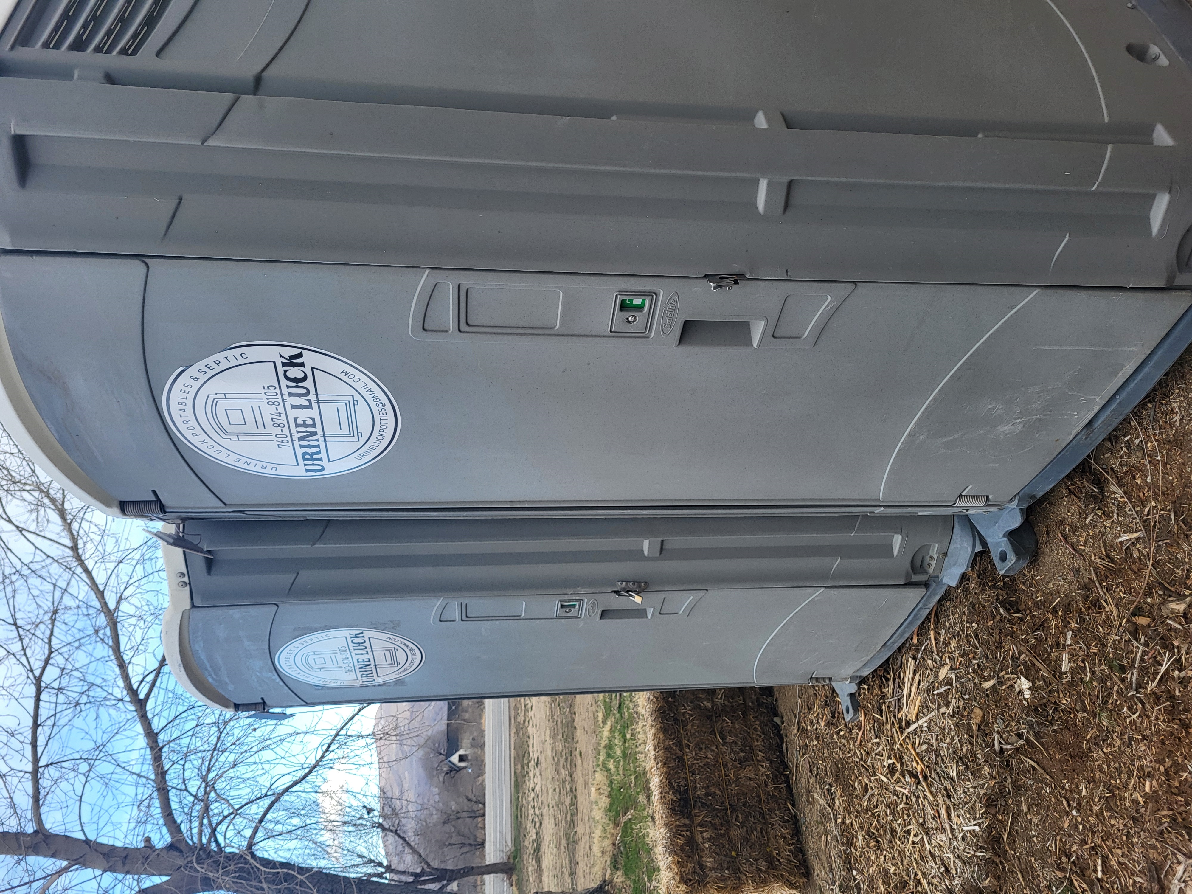 Two gray portable toilets side by side with Urine Luck company logos on their doors, placed outdoors on a dirt and mulch ground near trees and hay bales.