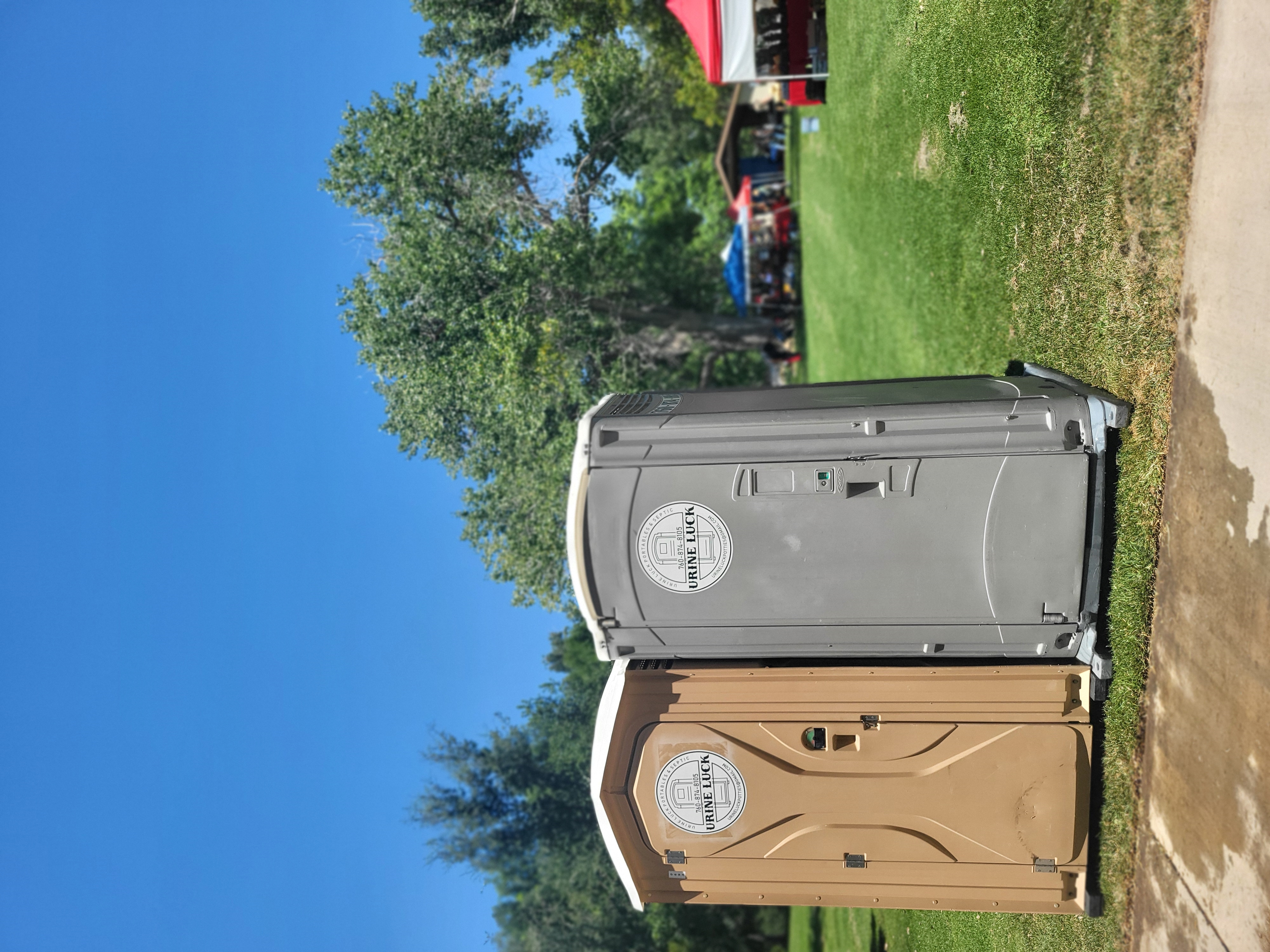 Two portable toilets, one brown and one gray, on grass in a park with tents and trees in the background under a clear blue sky.