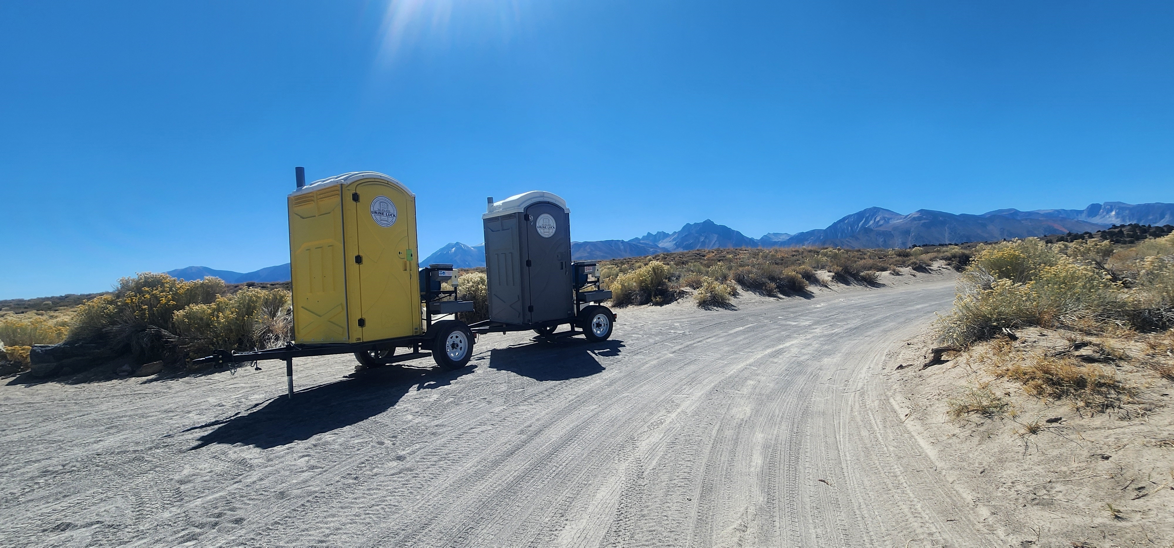 Yellow and gray portable toilets mounted on trailers parked on a dirt road in a desert landscape with mountains and clear blue sky in the background.