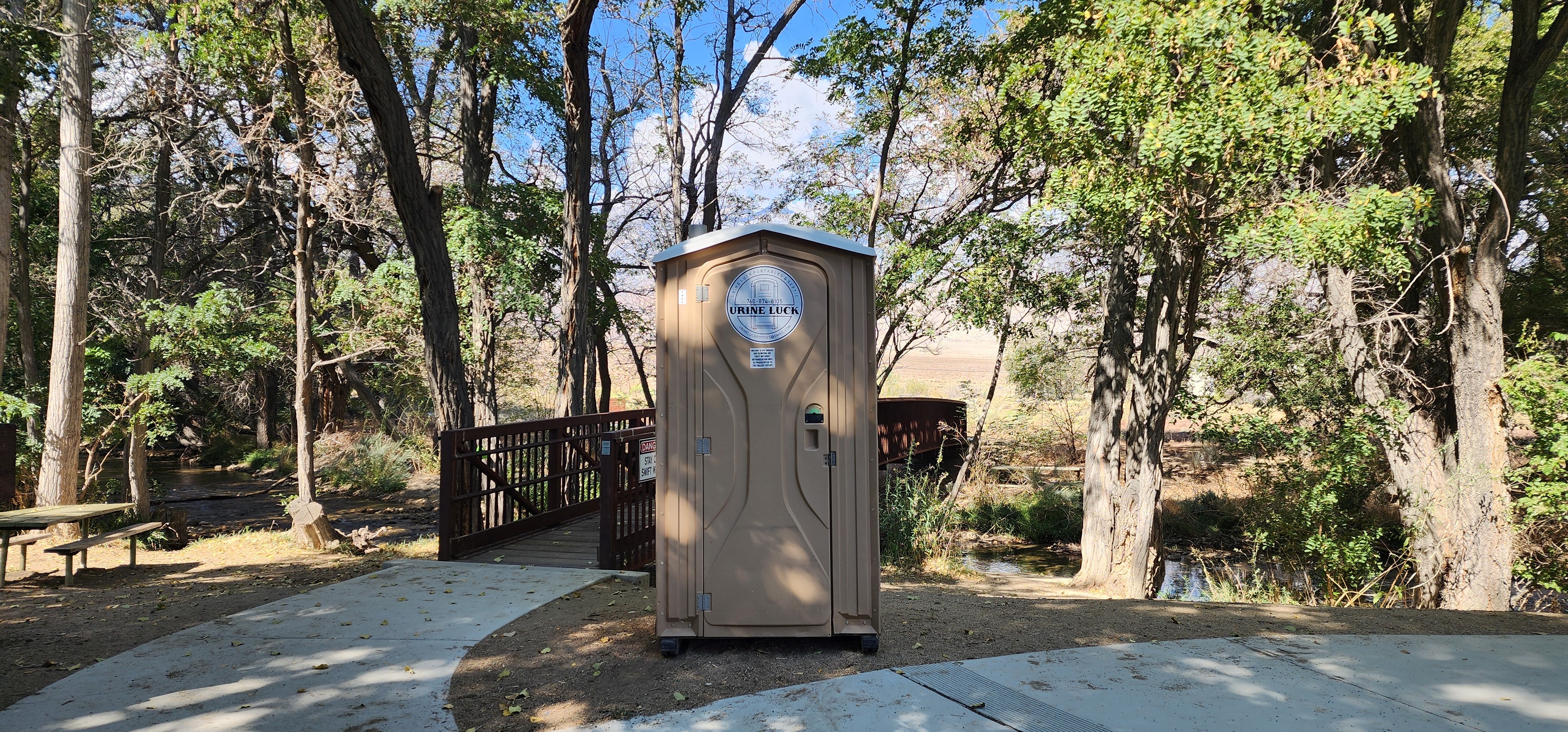 Portable restroom with 'Urine Luck' logo positioned on a dirt path near a small pedestrian bridge surrounded by trees and a creek.