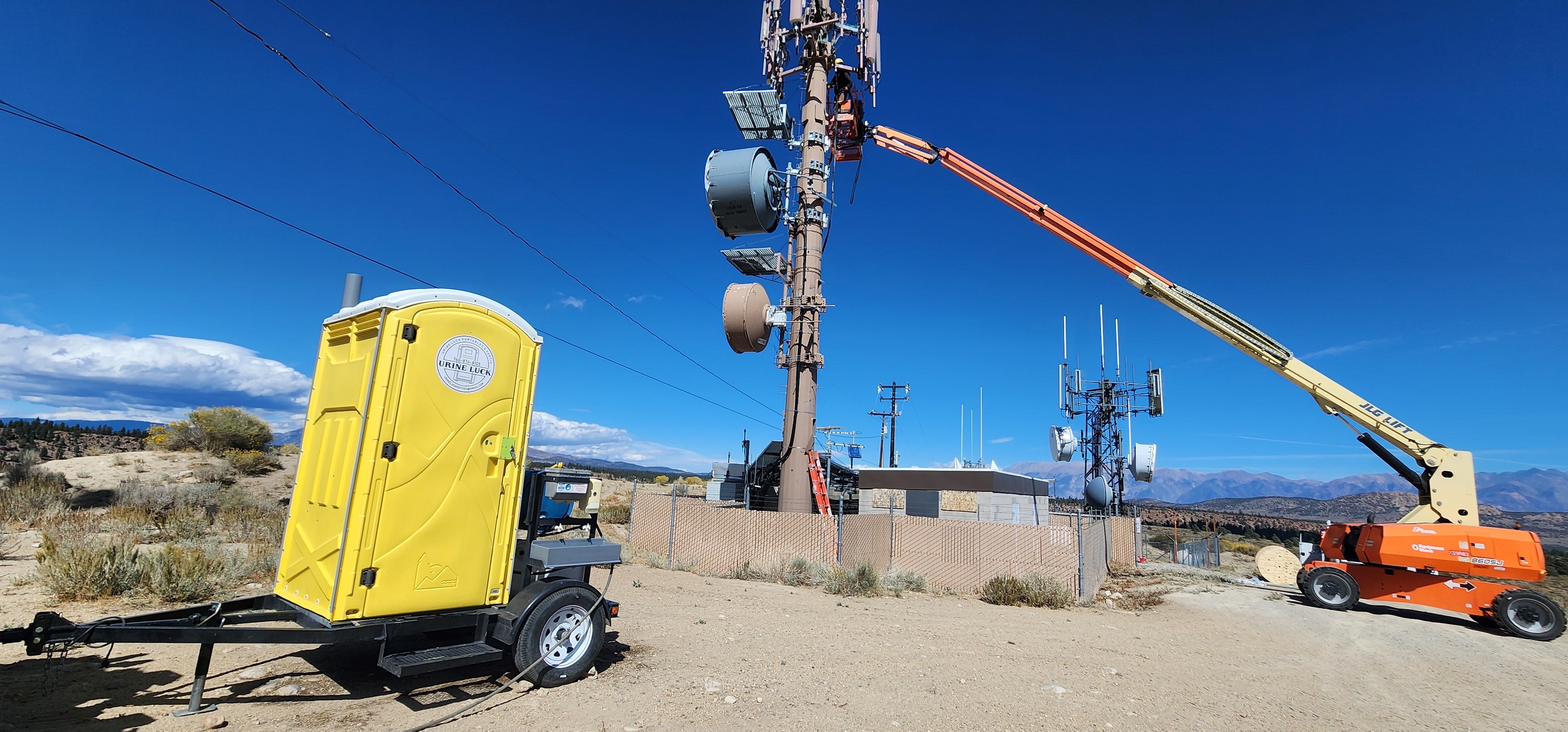 Yellow portable restroom on a trailer near a fenced telecommunications tower in a desert landscape under a clear blue sky.