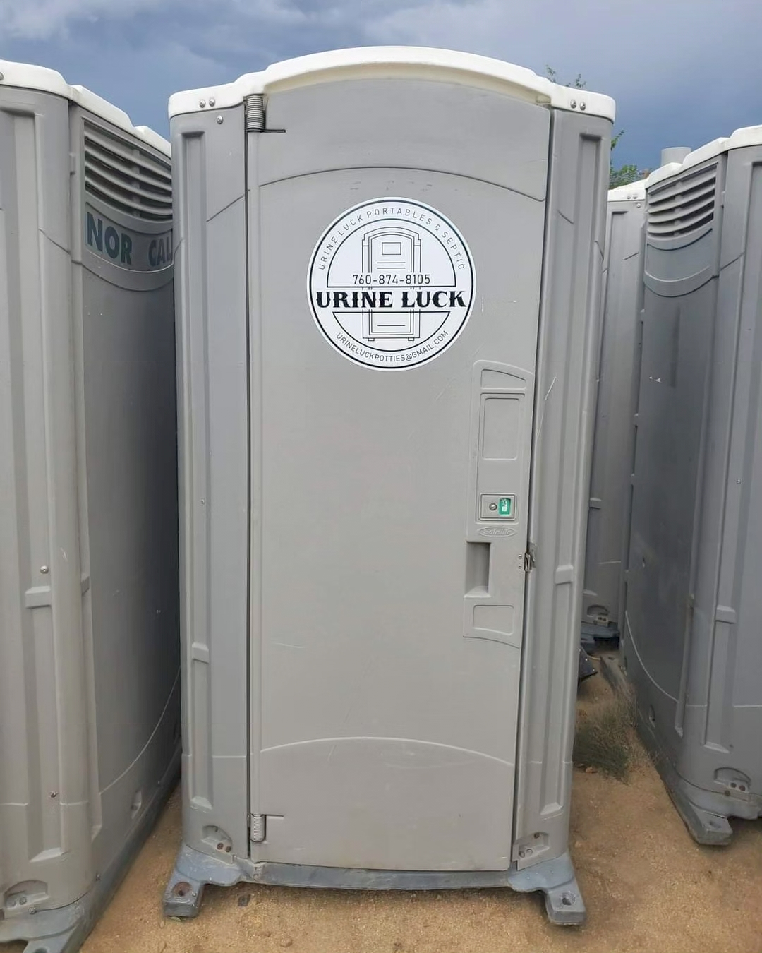 Gray portable restroom with a 'Urine Luck' logo and contact information on the door, placed on dirt ground between other portable restrooms.