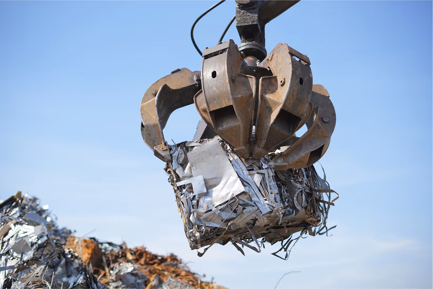 Industrial claw lifting crushed scrap metal against clear blue sky.