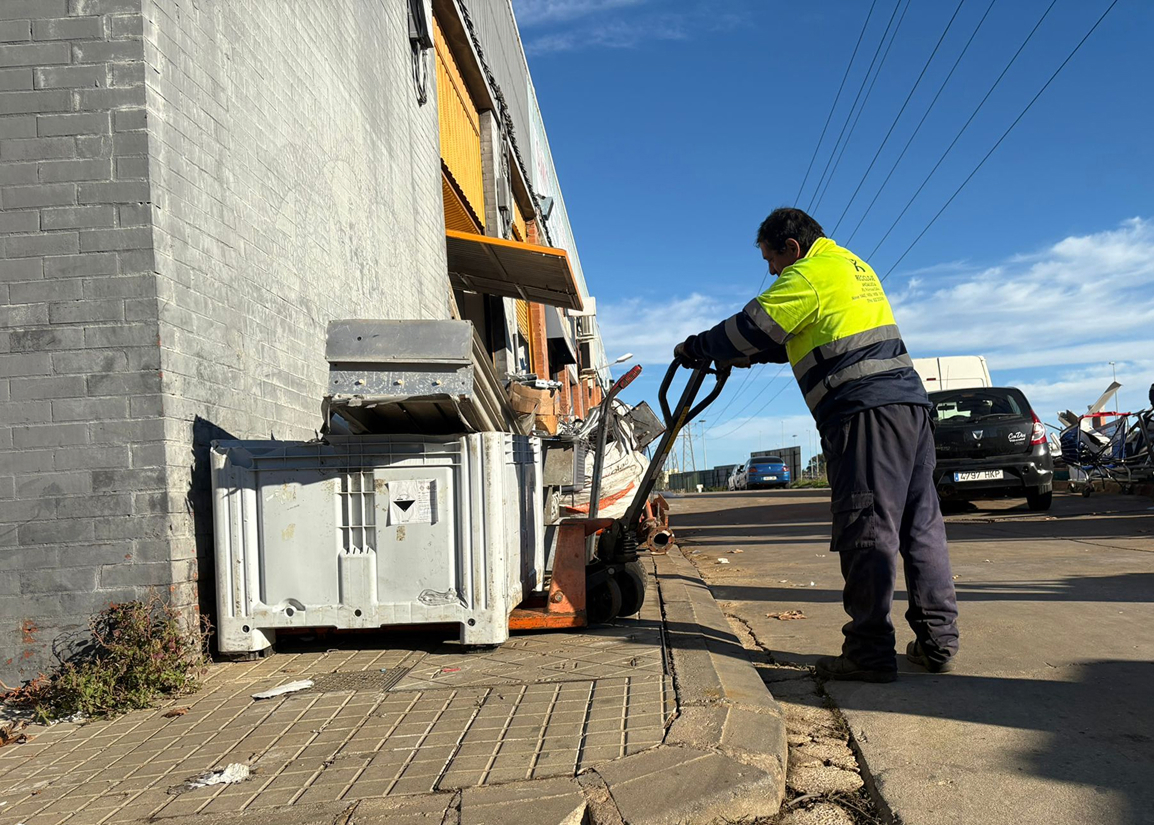 Worker in a yellow and navy uniform operating a pallet jack on a paved street next to a building with large bins during a sunny day.