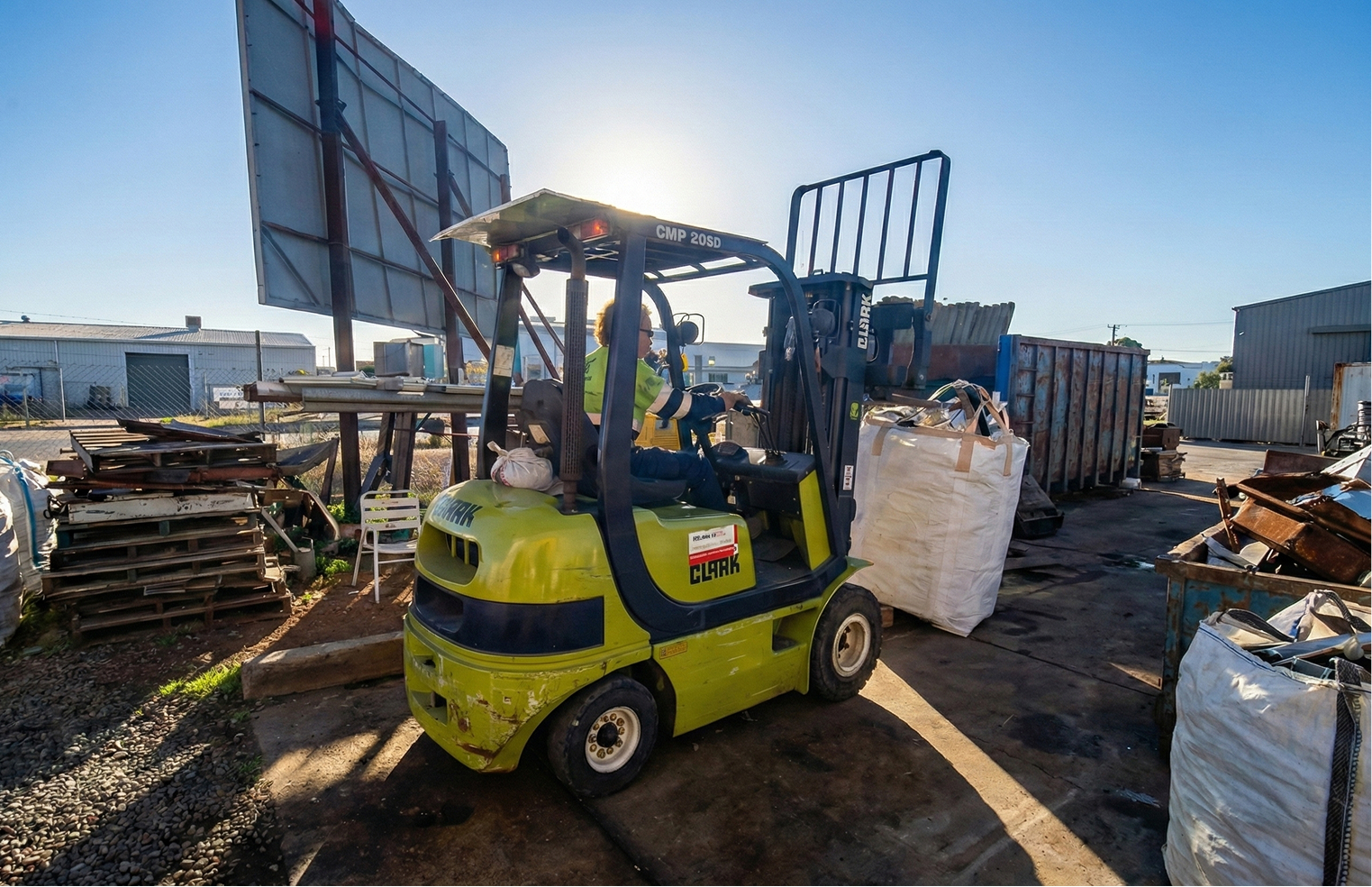 Worker operating a green Clark forklift in an industrial scrap yard during sunny daytime.